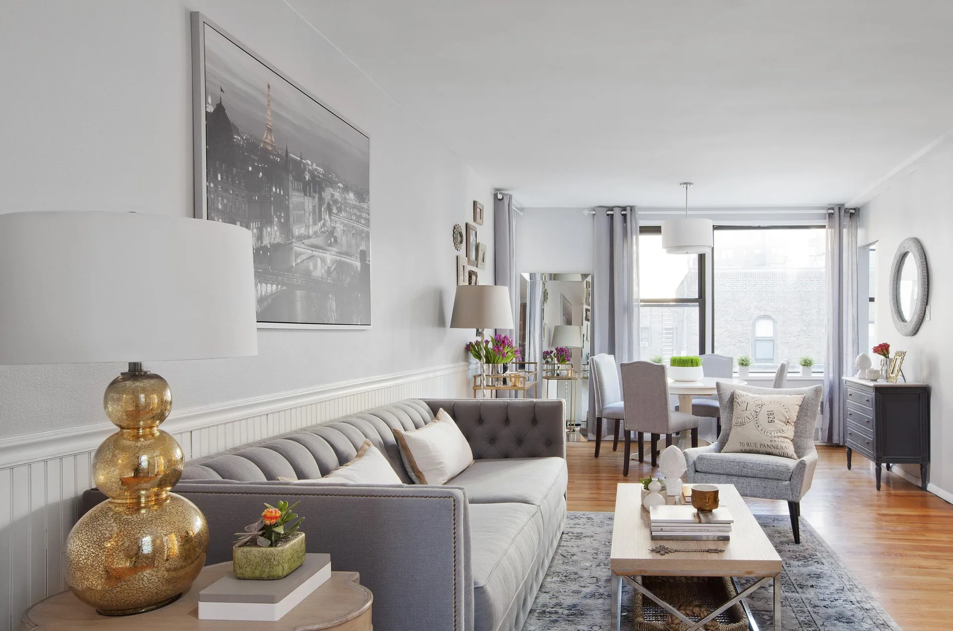 A grey tufted sofa sits in a living room featuring white beadboard wainscoting, a gold table lamp, and an adjacent dining area.