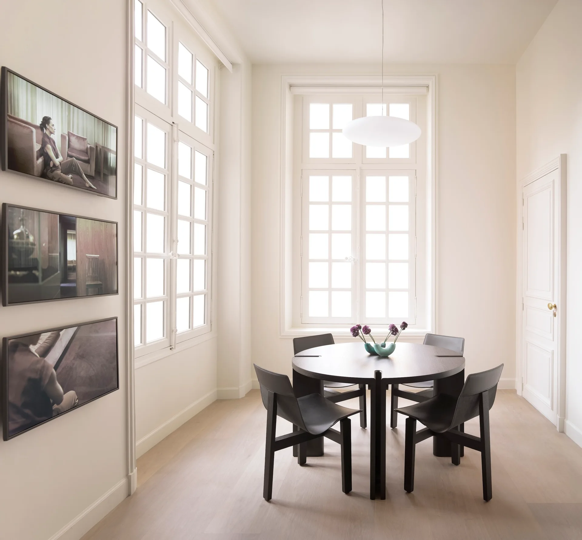 A minimalist dining room features a dark round wooden table with four black chairs, large white grid windows, and three framed artworks on the wall.