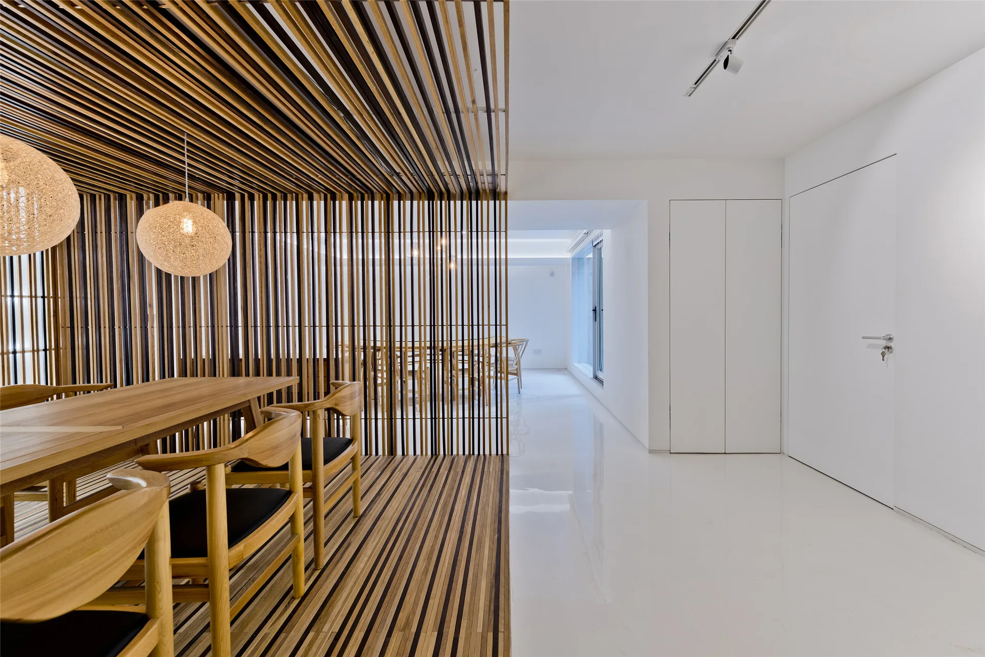 A minimalist dining room featuring a wooden table and chairs surrounded by a wooden slat floor, wall, and ceiling divider next to glossy white flooring.