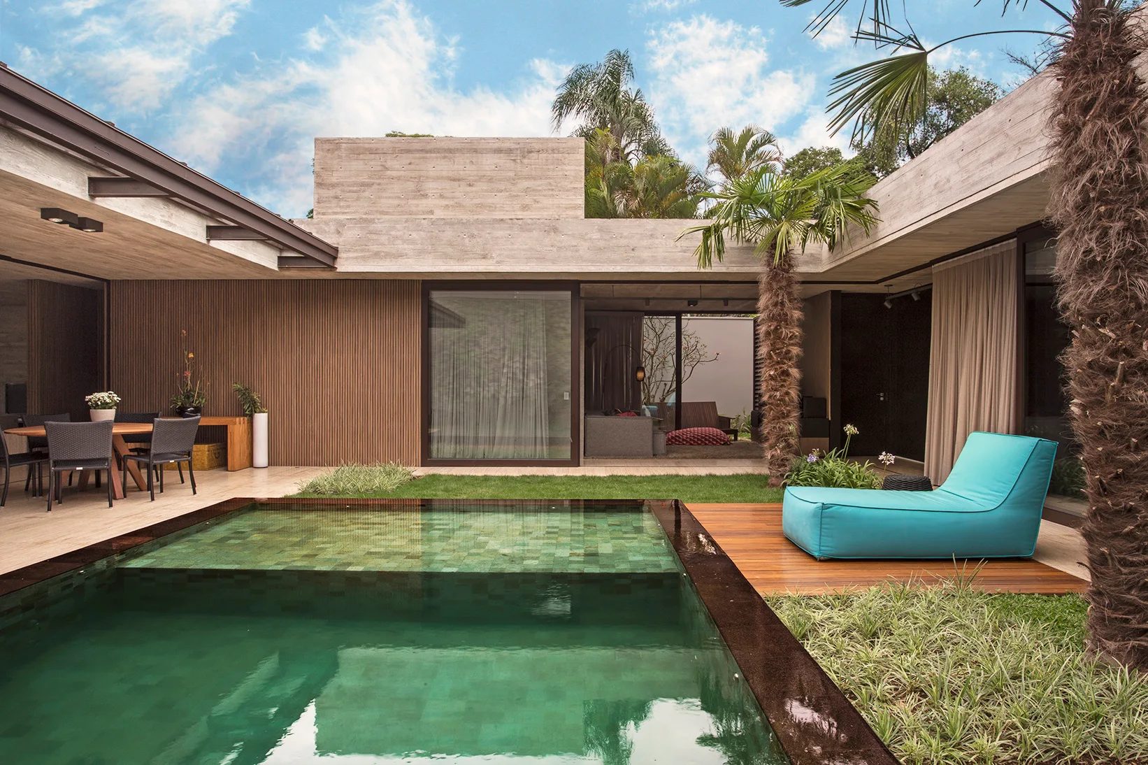A contemporary backyard pool is bordered by a wooden deck with a turquoise lounge chair and a patio sheltered by an exposed concrete roof.