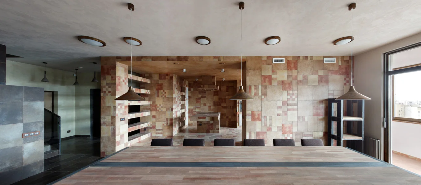 A large wooden dining table with dark chairs sits in front of a multi-colored earth-tone tiled feature wall with three pendant lights overhead.