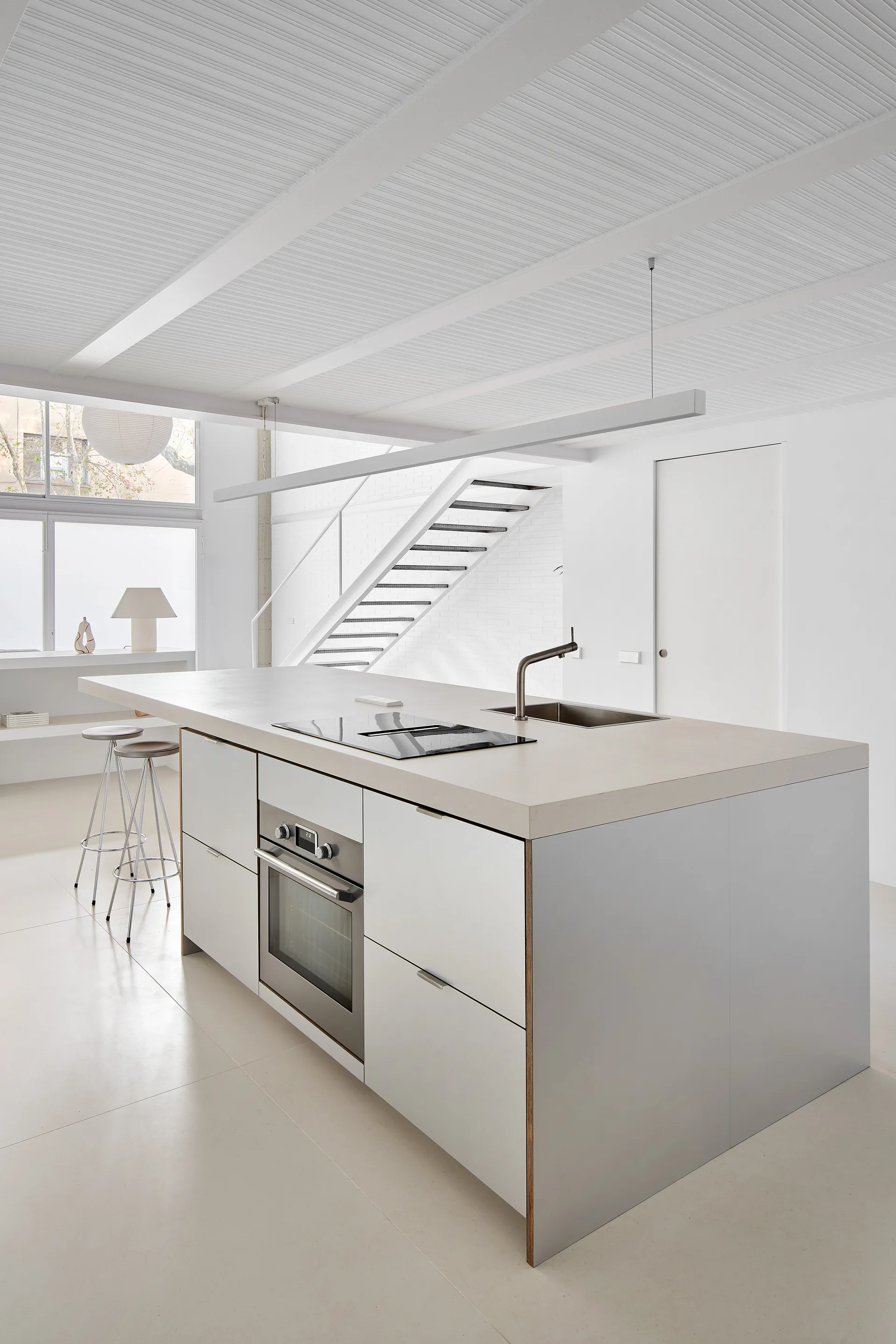 A minimalist white kitchen featuring a large central island with a built-in oven, sink, and cooktop, with a staircase visible in the background.