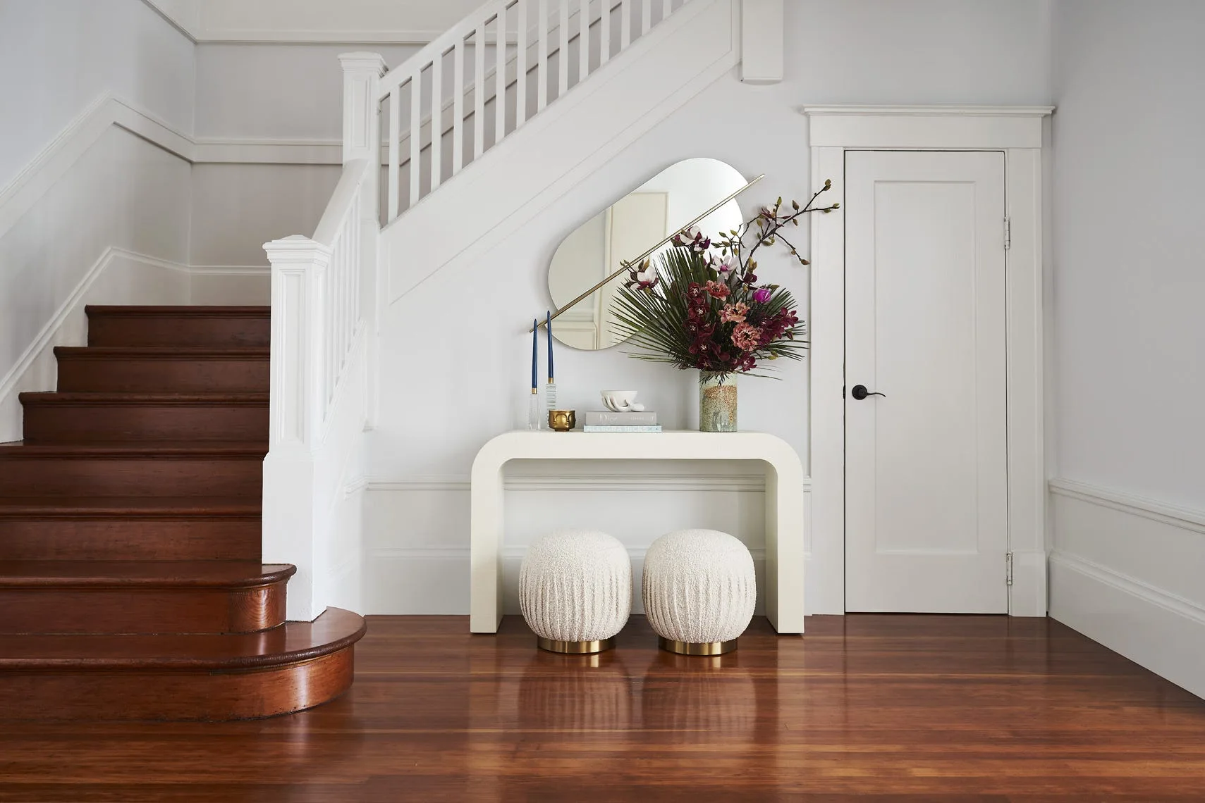 A curved white console table with two round ottomans underneath is positioned next to a wooden staircase in an entryway.