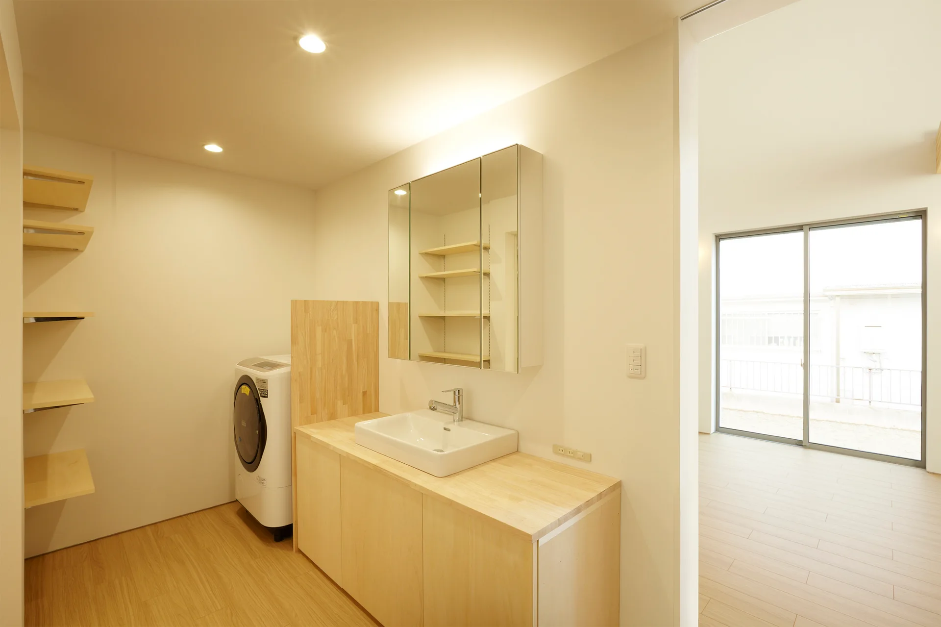A minimalist laundry and vanity room featuring a light wood washbasin cabinet, a washing machine, open wooden shelves, and a mirrored medicine cabinet.