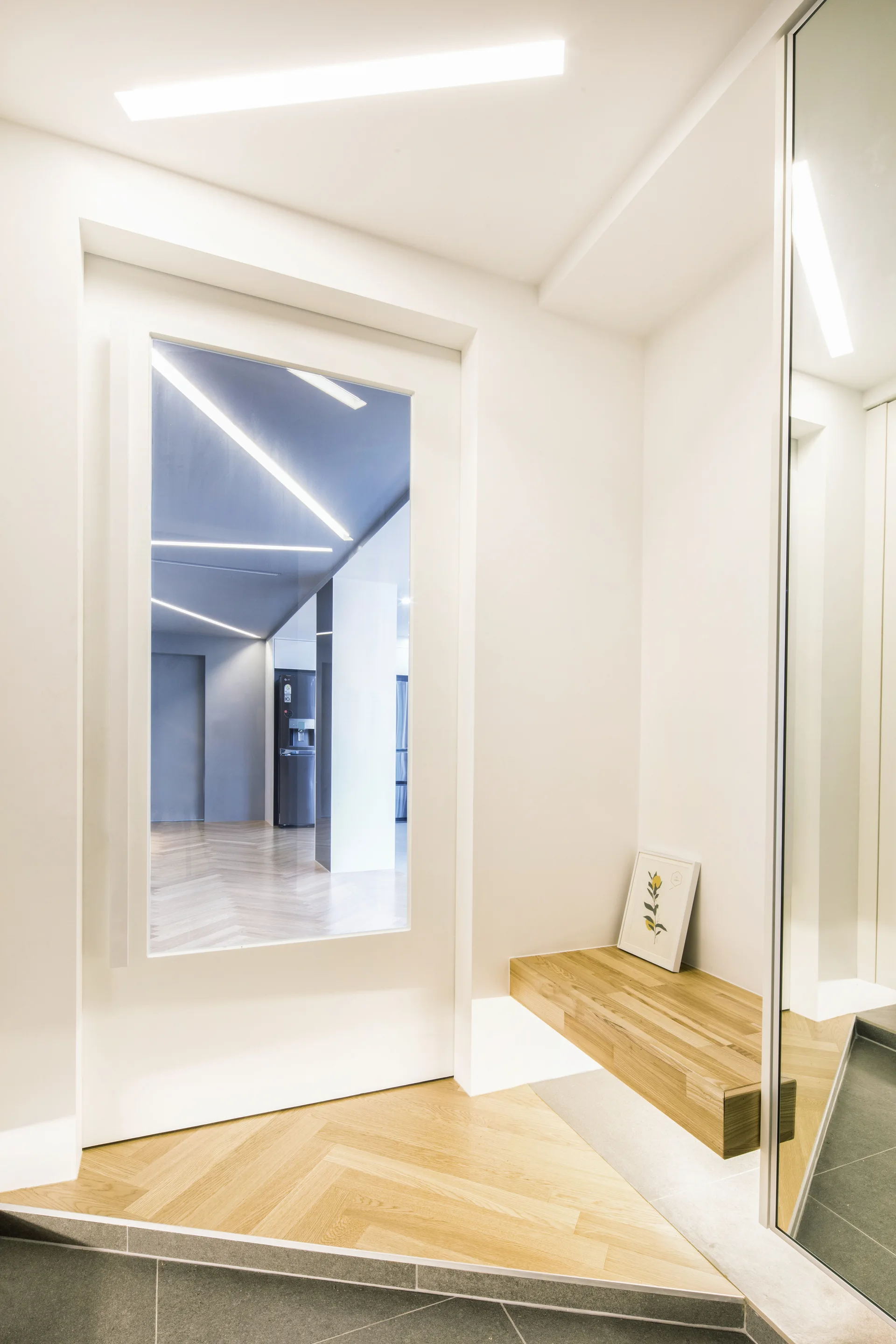A minimalist white entryway featuring a floating light wood bench, herringbone parquet flooring, and a large glass-paneled door.