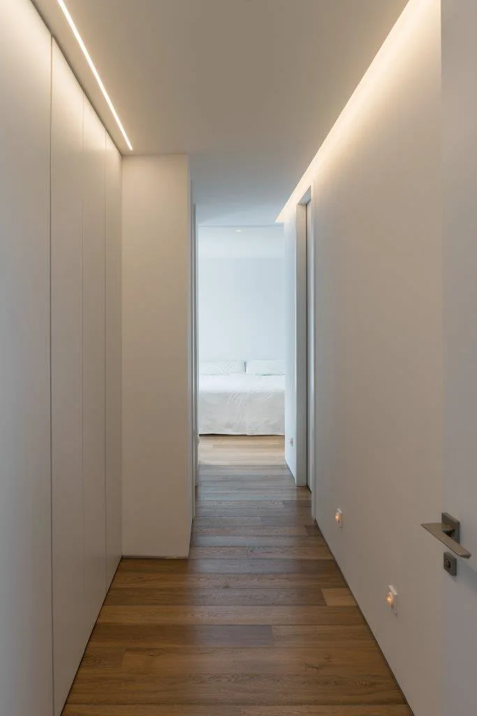 A minimalist white hallway featuring built-in cabinets, warm plank wood flooring, and integrated ceiling cove lighting leads to a distant bedroom.