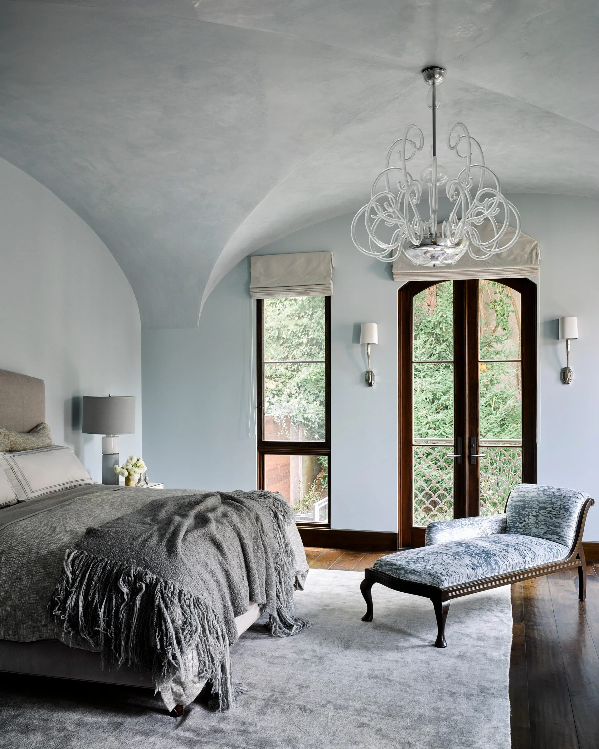 A pale blue bedroom featuring a grey upholstered bed, a curved glass chandelier hanging from a vaulted ceiling, and a patterned chaise longue.