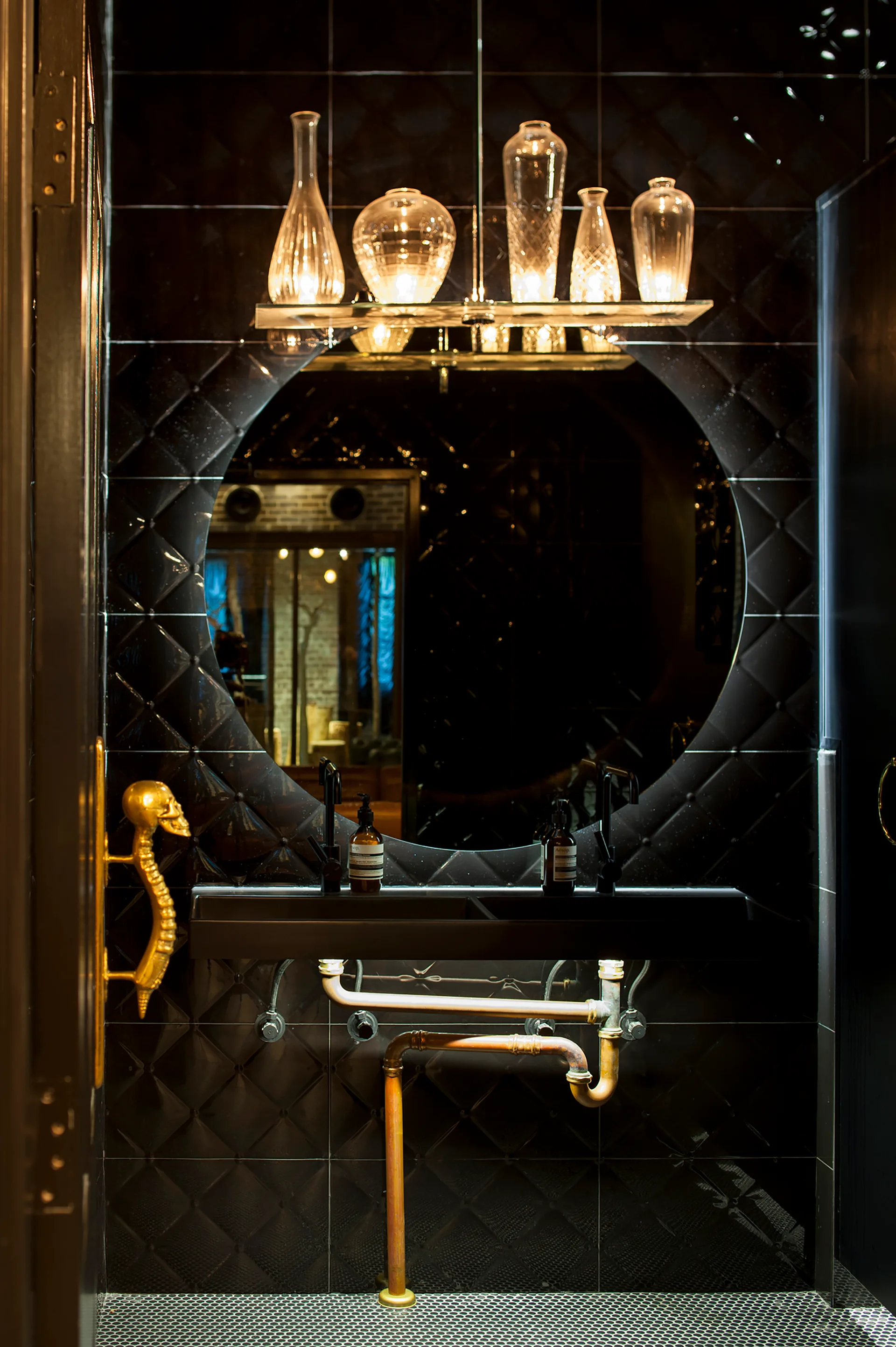 A dark powder room features glossy black quilted wall tiles, a round mirror, a floating sink with exposed copper pipes, and a gold skull door handle.