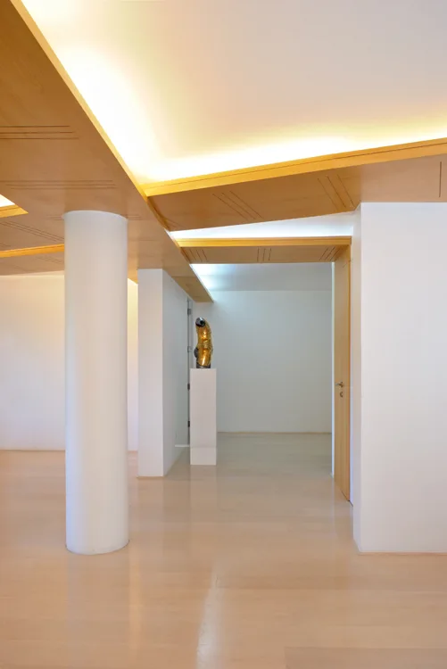 A minimalist white room features a cylindrical support column, light wood flooring, and a geometric wood-paneled ceiling with warm glowing cove lights.