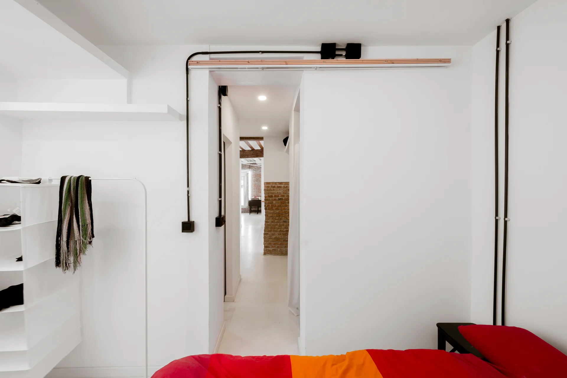 A minimalist white bedroom featuring exposed black wiring conduits on the walls and a bed with vibrant red and orange bedding.