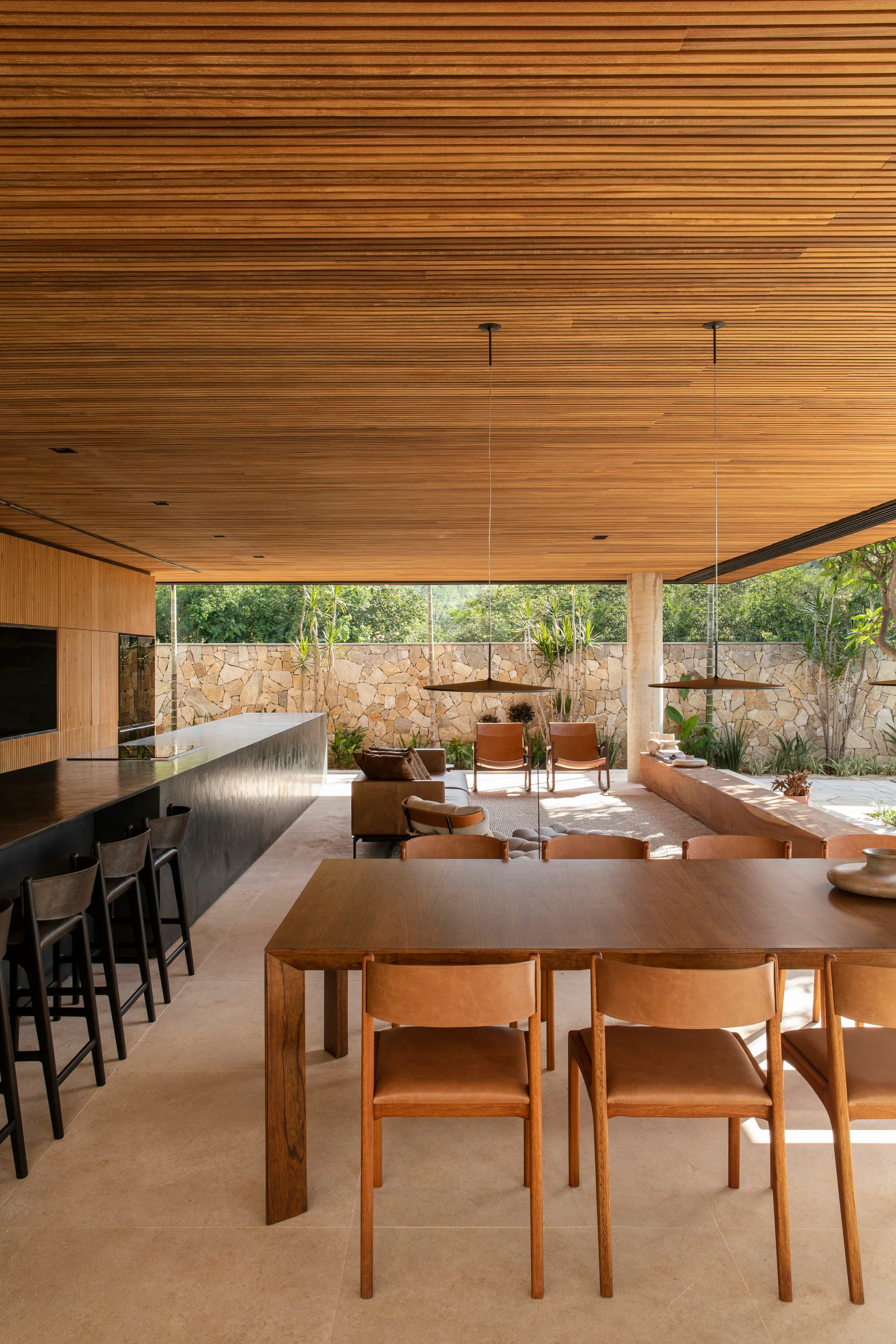 An open-concept dining and living area features a wooden table, leather chairs, a long black kitchen island, and a continuous slatted wood ceiling.