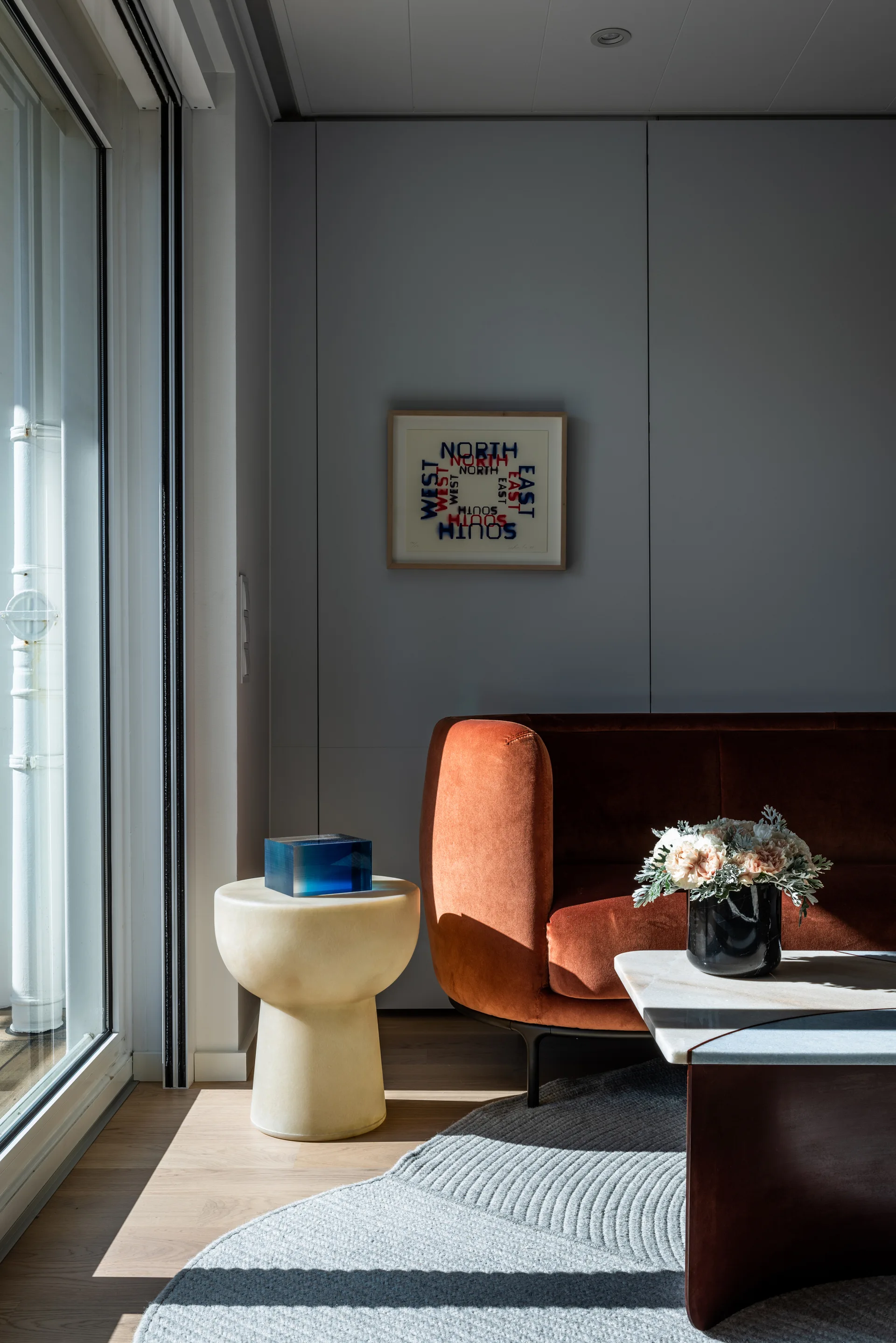 A rust-colored velvet sofa sits beside a cream sculptural side table and a grey textured rug under natural sunlight from a large window.