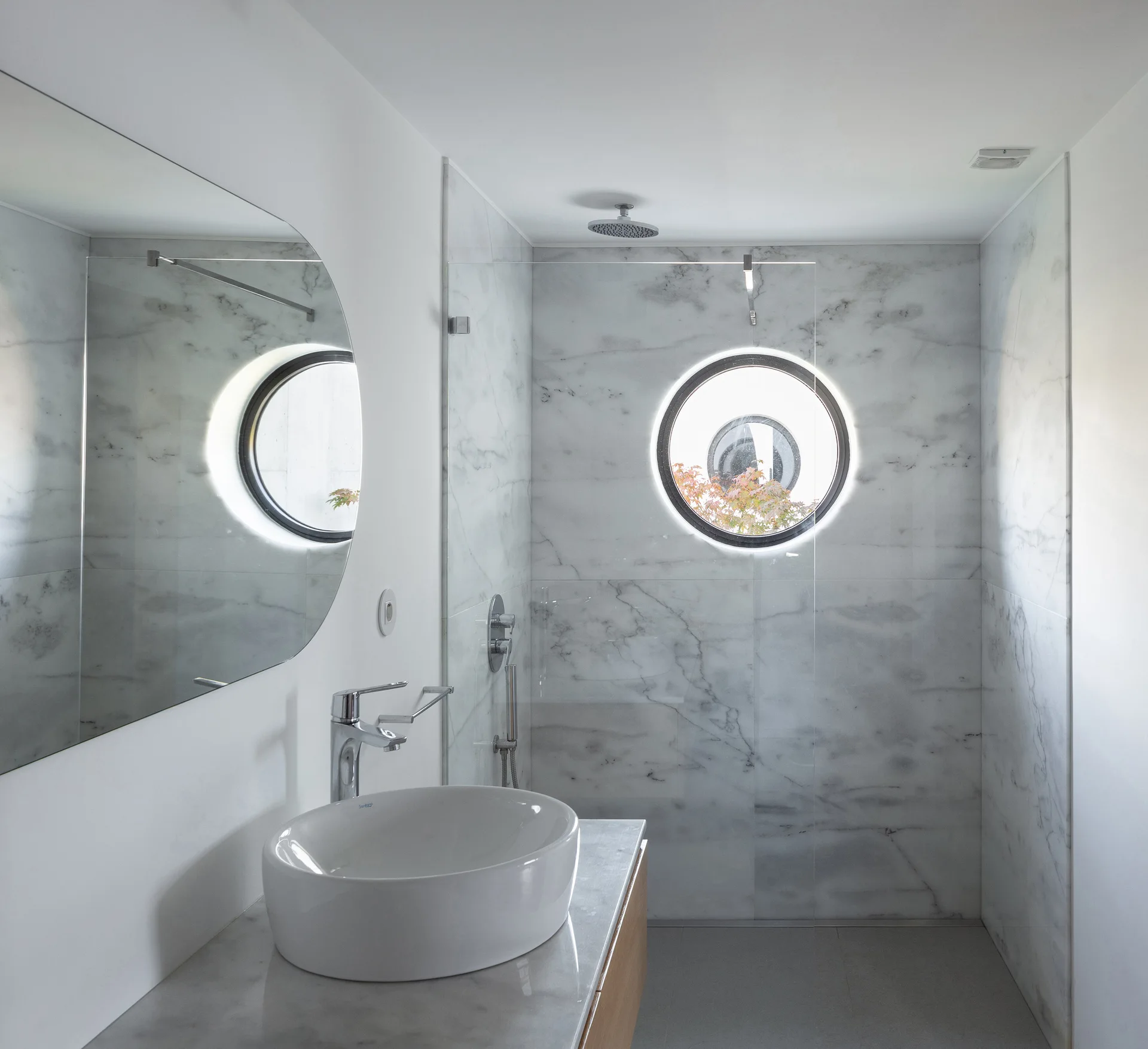 A minimalist bathroom featuring a white vessel sink on a marble countertop and a walk-in marble shower with a round porthole window.