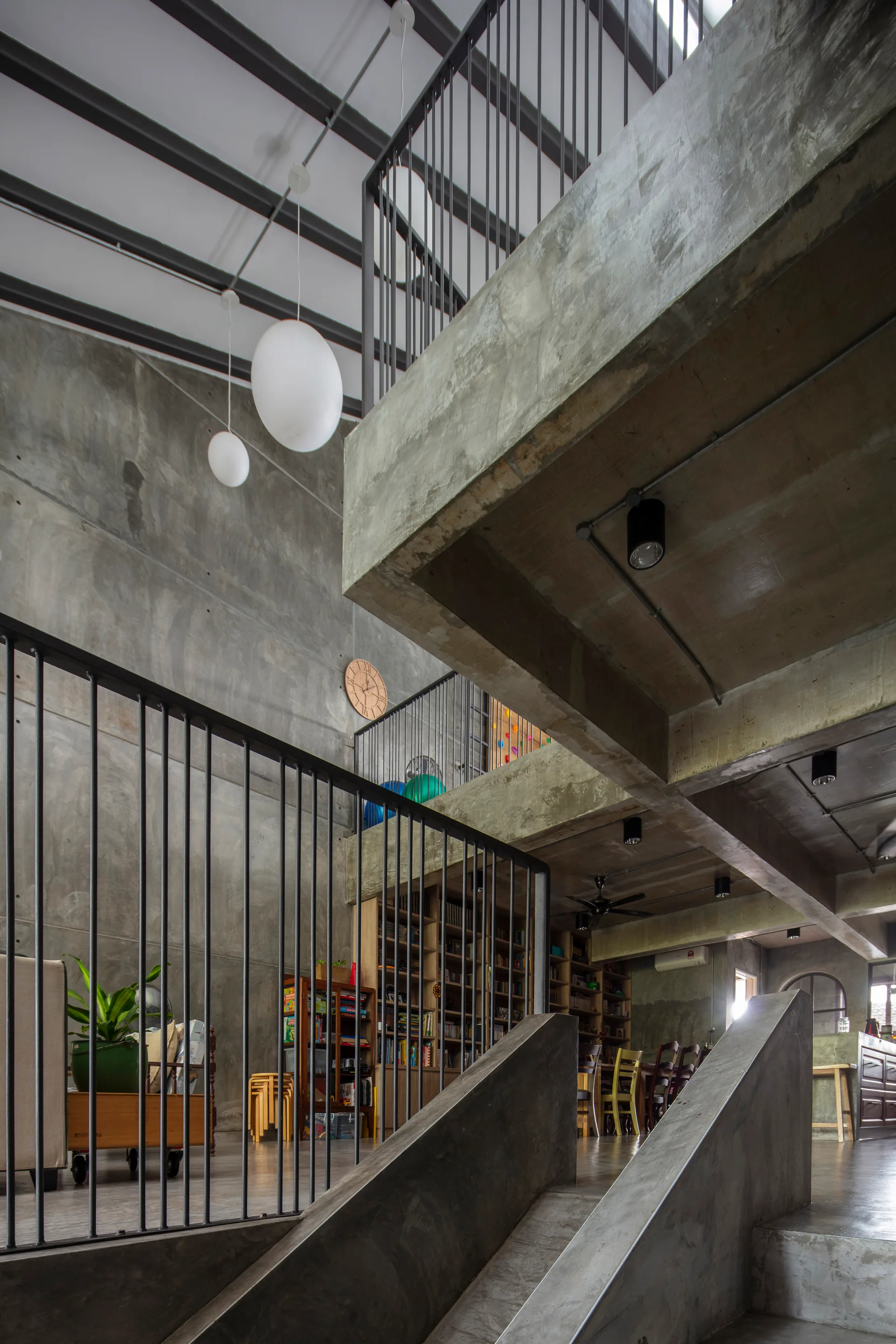 A multi level industrial interior features raw concrete walls, a concrete staircase, black vertical metal railings, and hanging globe pendant lights.