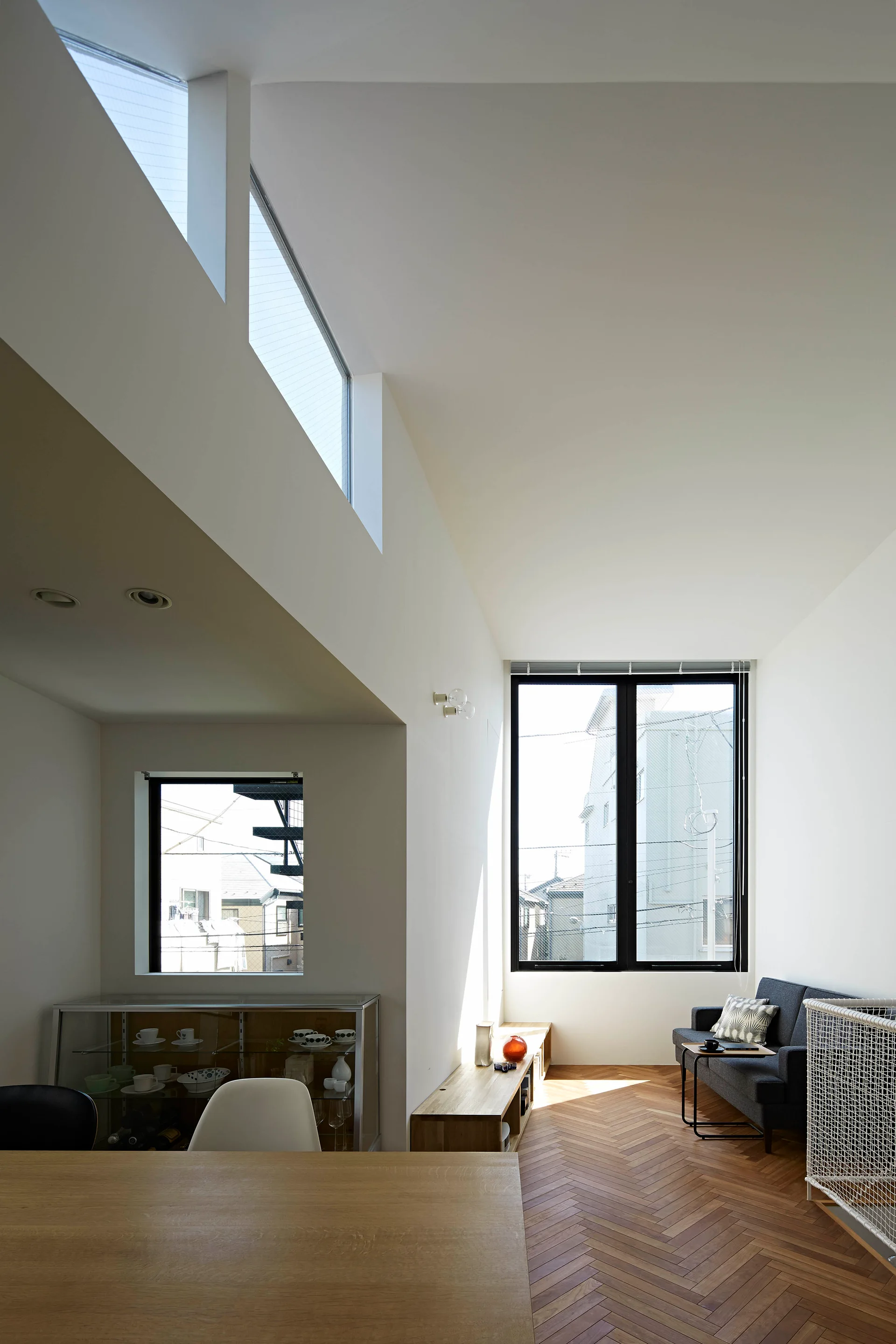 A minimalist living room features herringbone wood flooring, a low wooden media console, a grey sofa, and high clerestory windows on white walls.