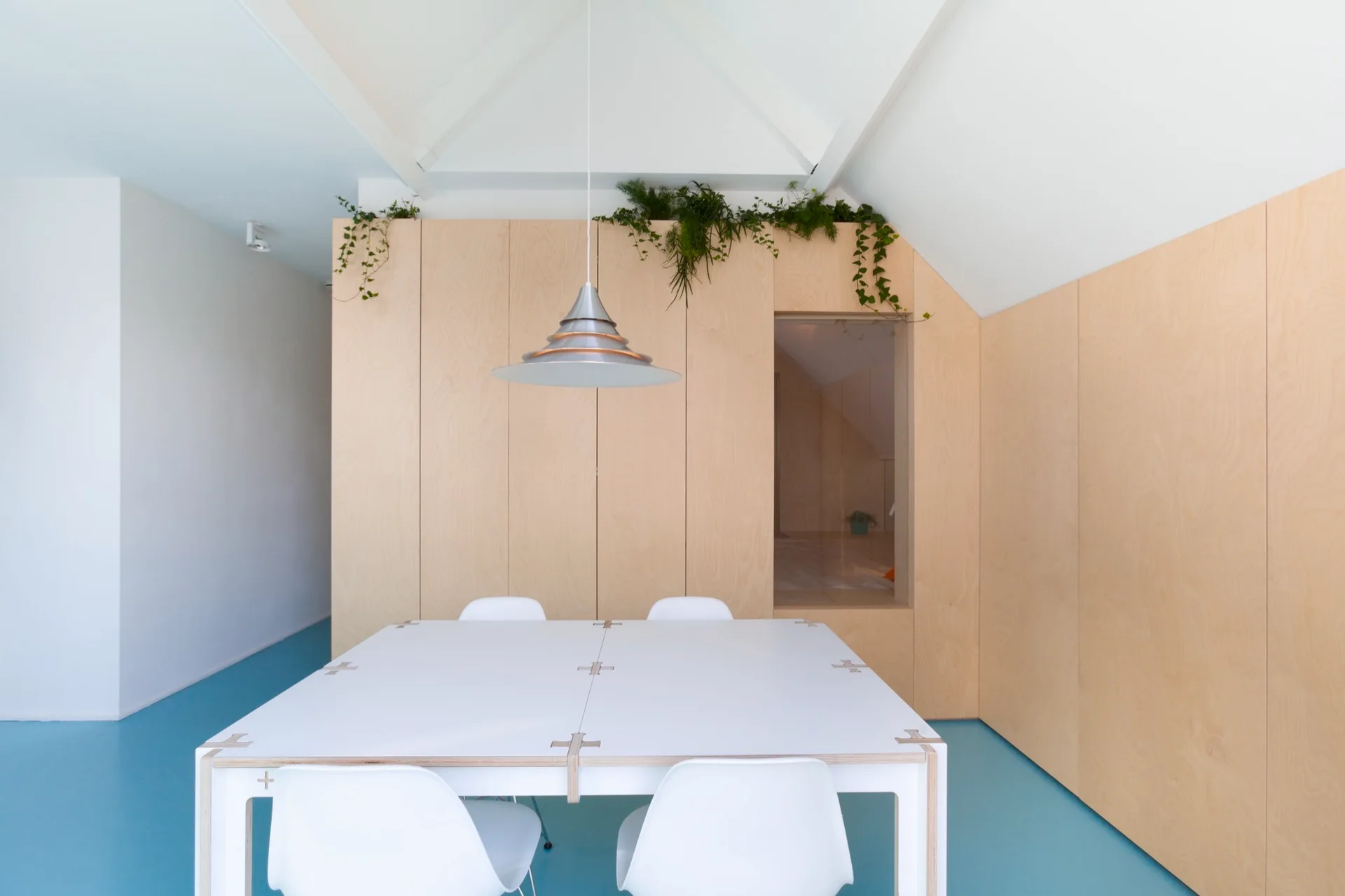 A minimalist dining room features a white square table and chairs on a bright blue floor beneath a light wood paneled wall and vaulted ceiling.