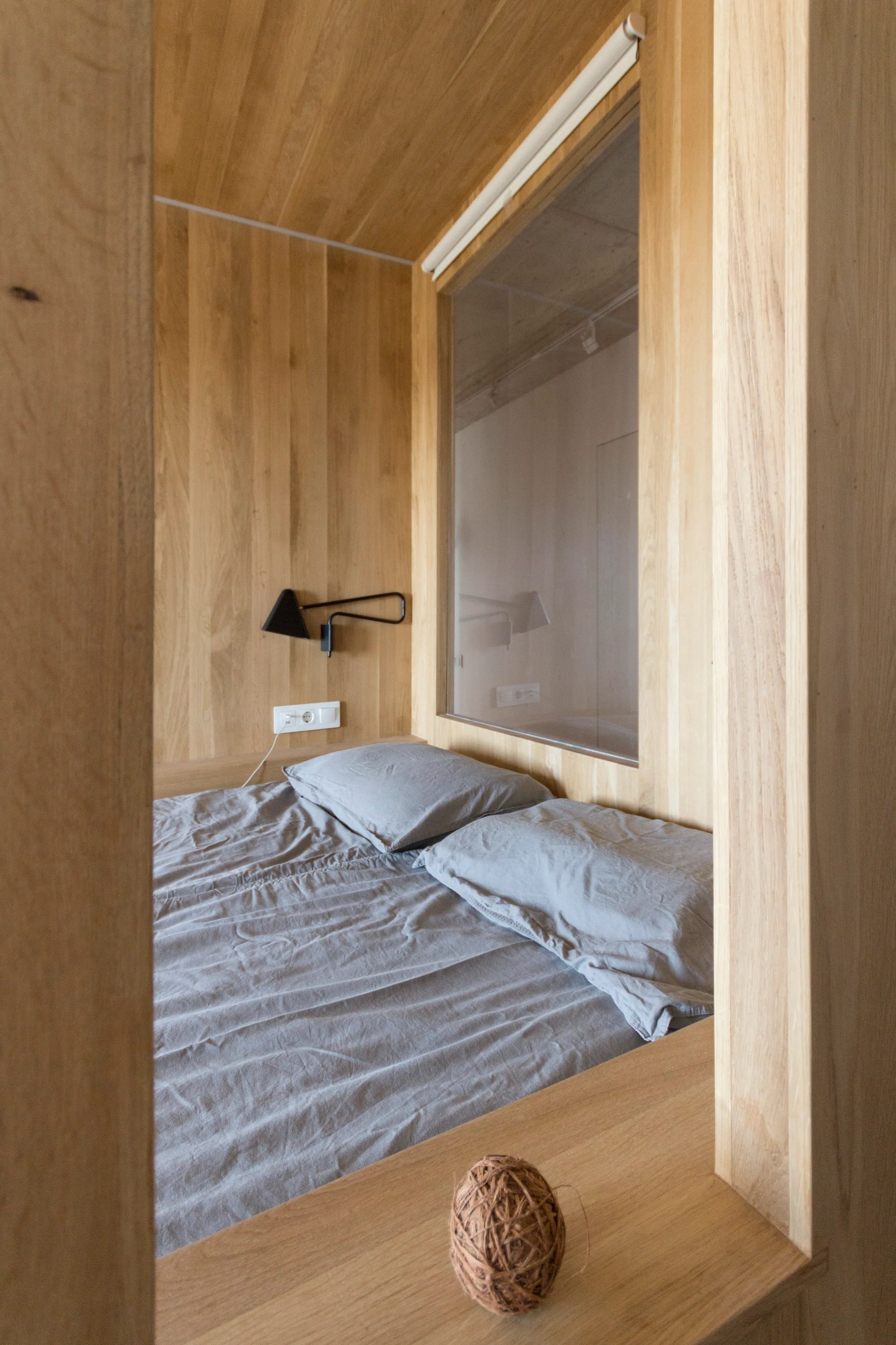 A minimalist wood-paneled sleeping alcove features a bed with grey linens, a black wall lamp, and a large glass window pane.