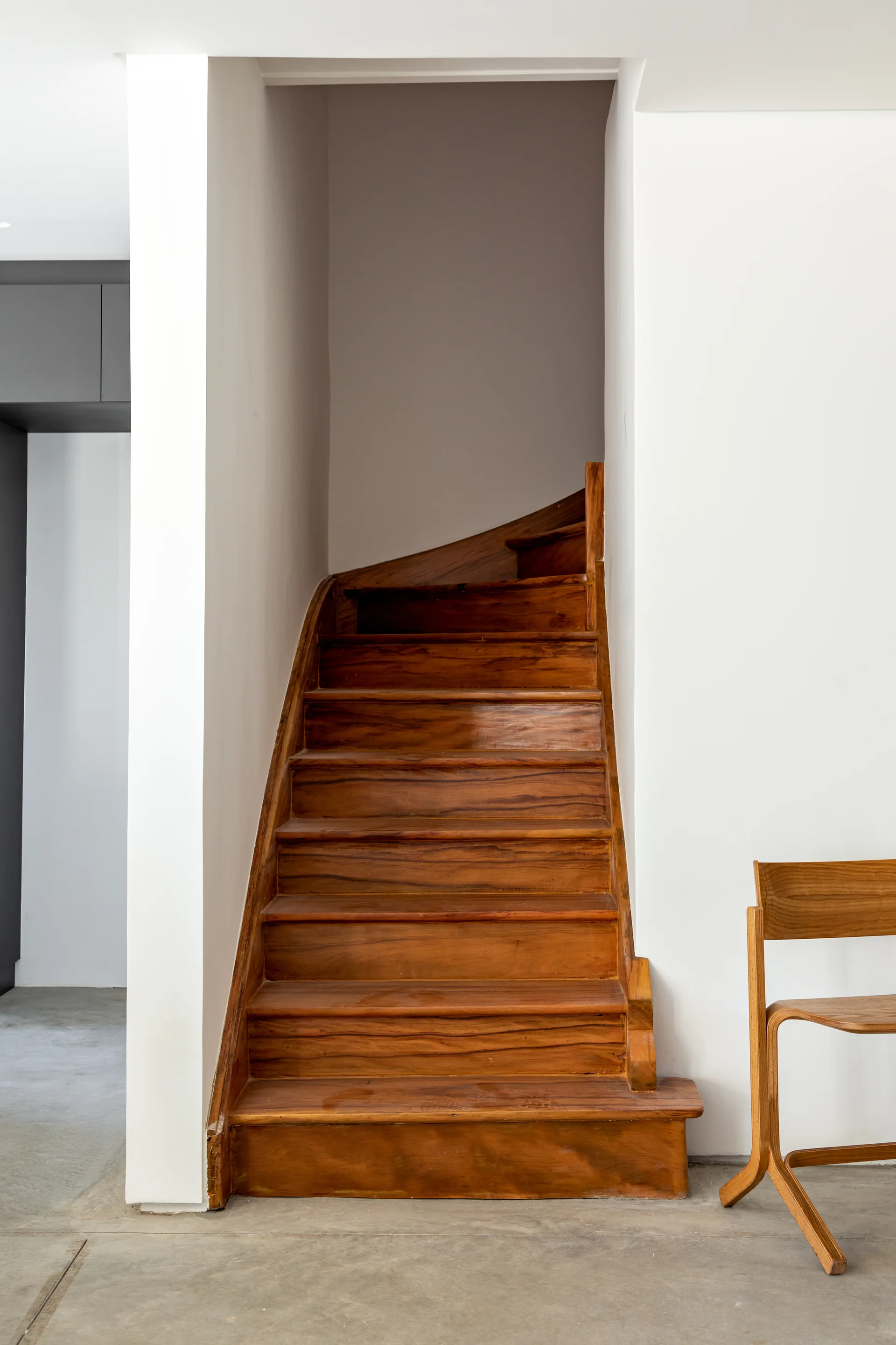 A warm wood staircase curves upward between two smooth white walls next to a modern wood chair on a grey concrete floor.