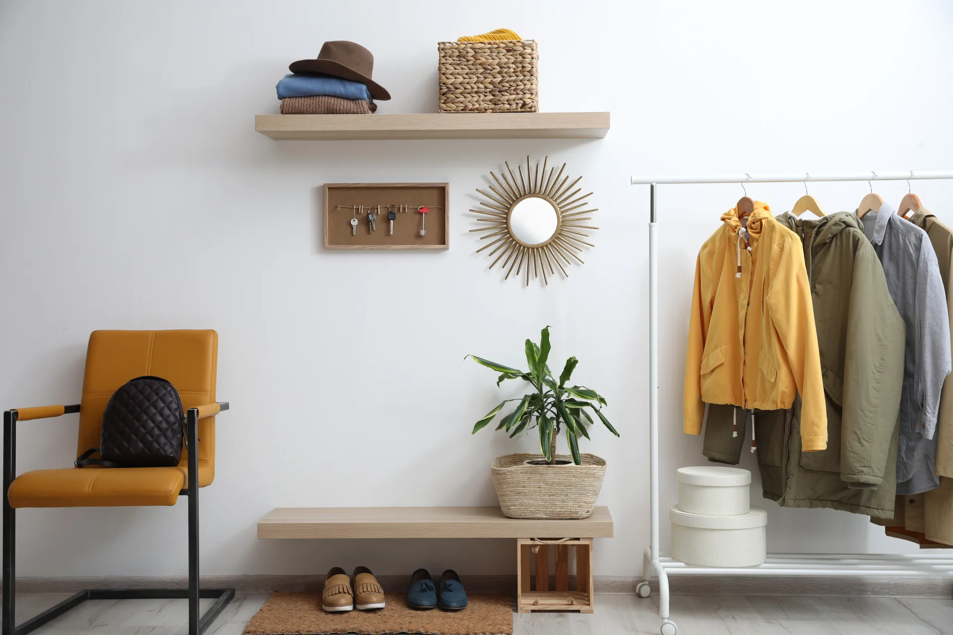 An organized entryway with a white clothing rack, mustard yellow chair, floating wood shelves, and a starburst mirror on a white wall.