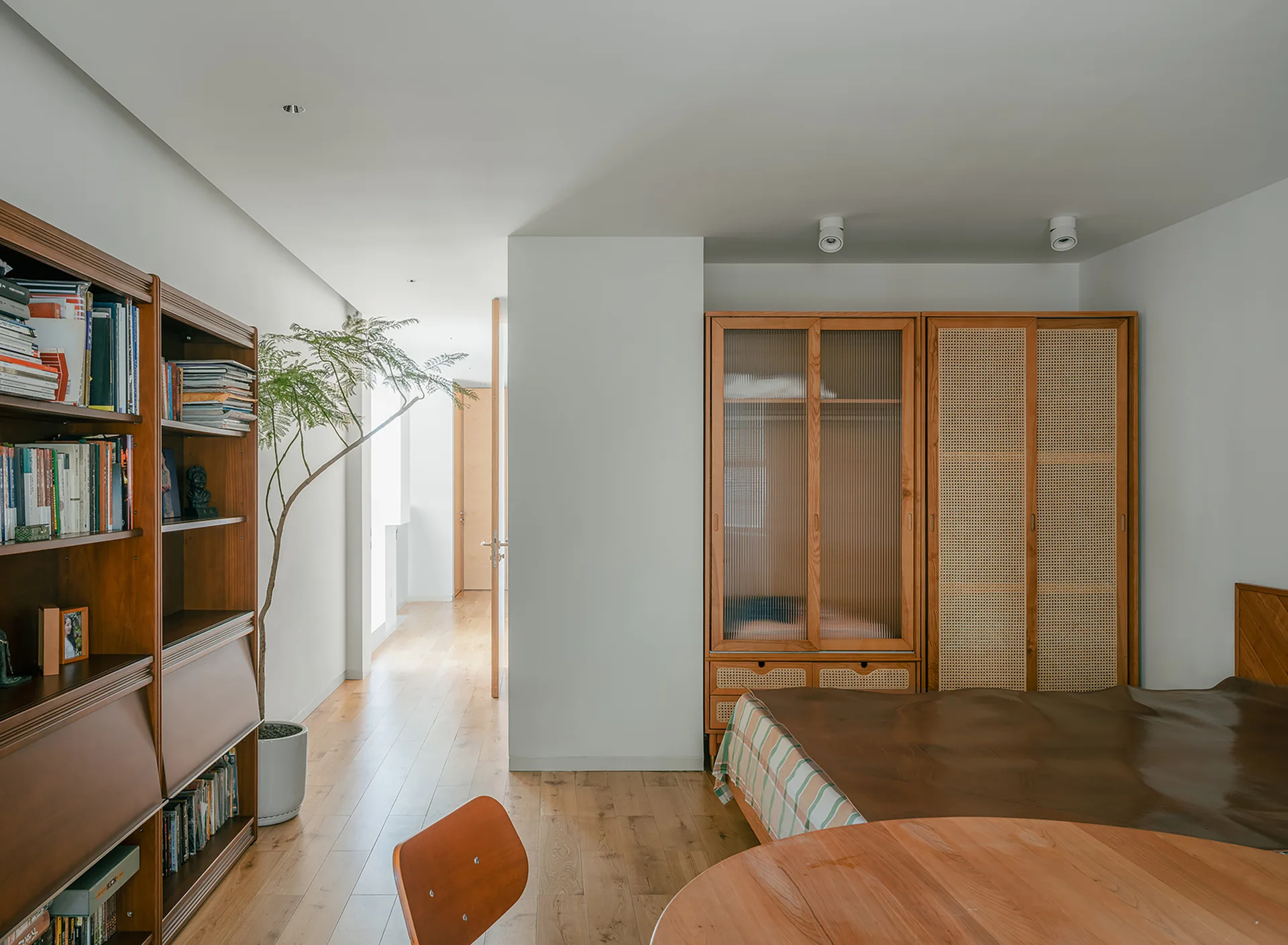 A minimalist bedroom features a wooden wardrobe with rattan and fluted glass doors, white walls, and a tall bookcase.