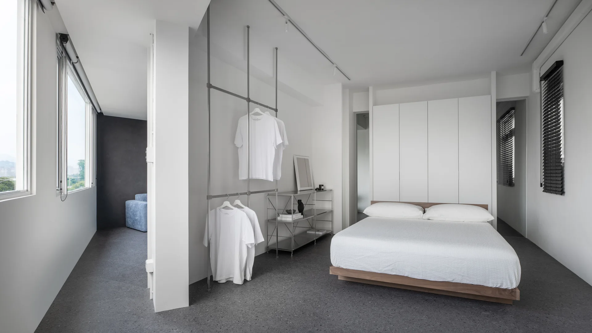A minimalist white bedroom features a wood-base platform bed, integrated tall cabinets, and an open metal clothing rack on dark speckled flooring.