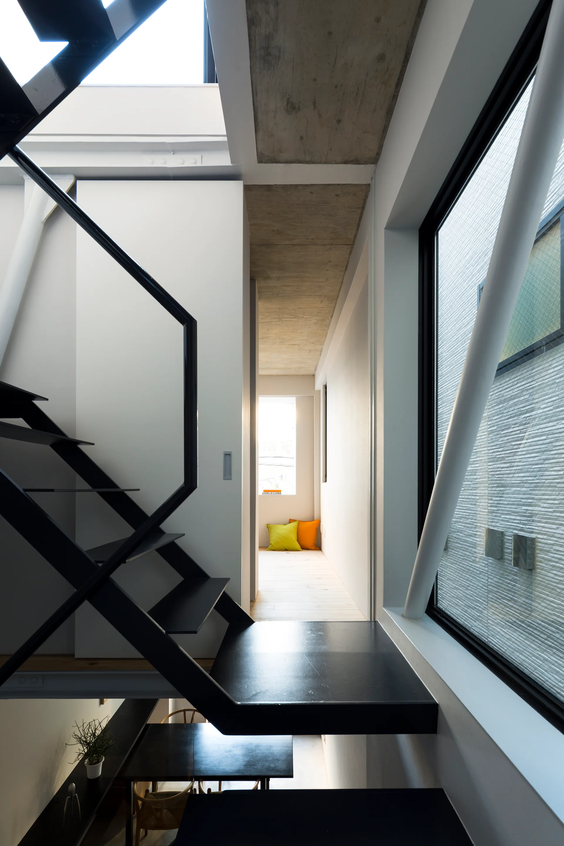 A matte black open-tread metal staircase sits adjacent to white walls, a large window, and an exposed raw concrete ceiling.