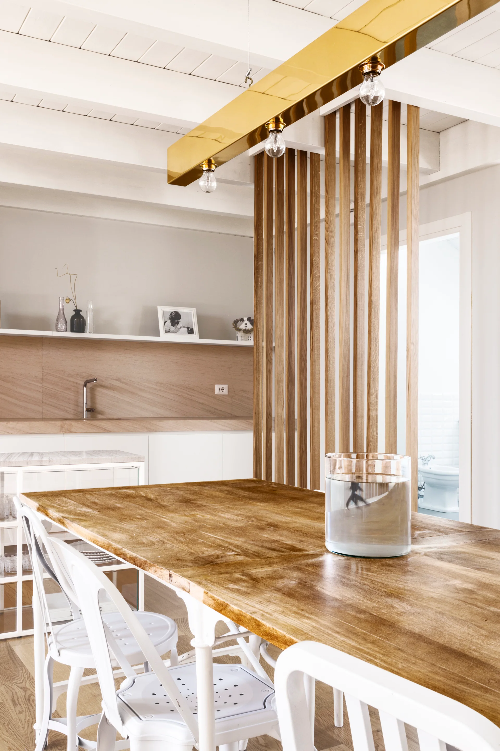 A wooden dining table with white metal chairs is separated from a kitchen by a vertical slatted wood screen under painted ceiling beams.