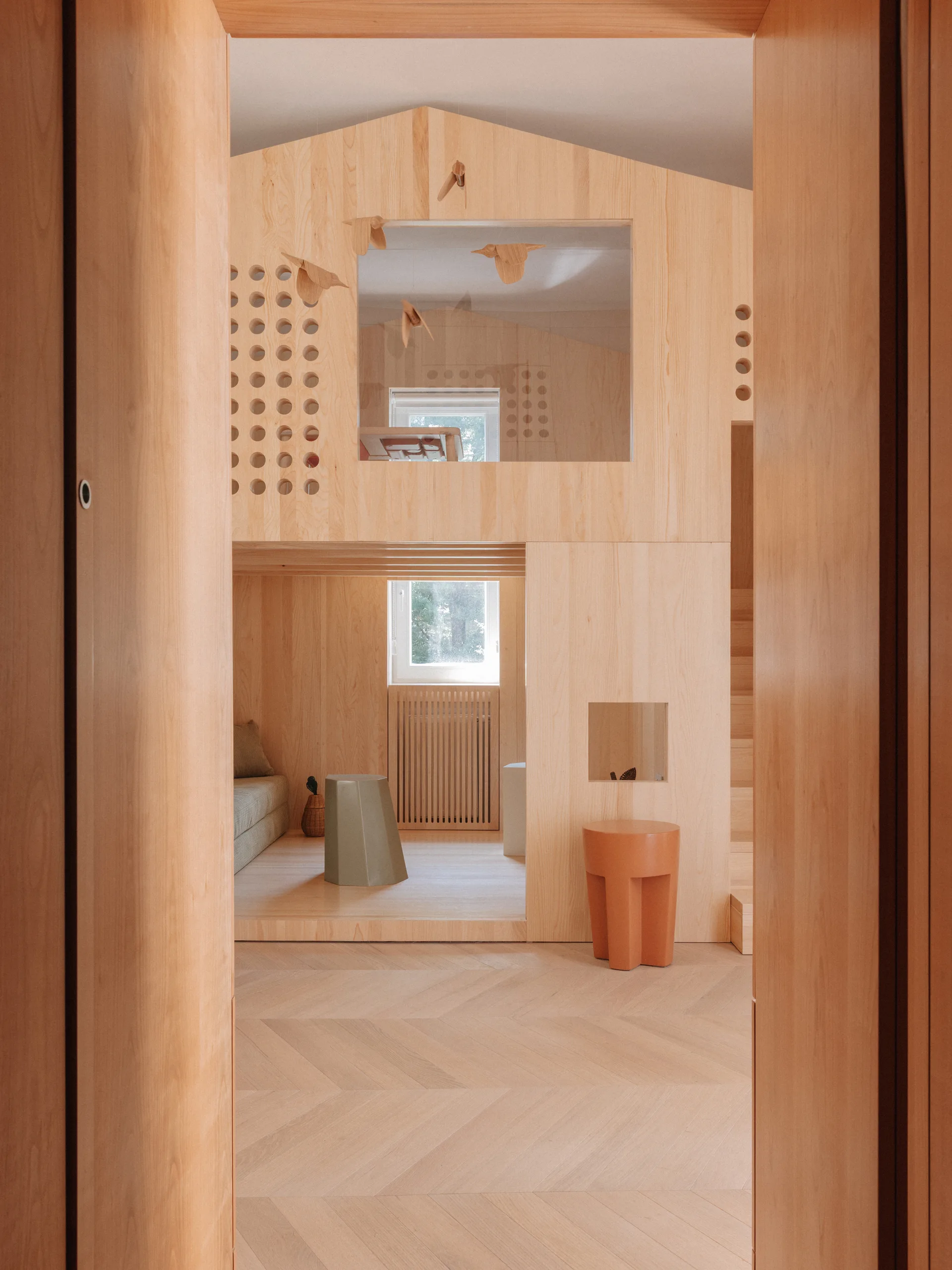 A kids playroom features a custom wooden playhouse with an integrated loft, circular cutouts, and stairs on herringbone flooring.