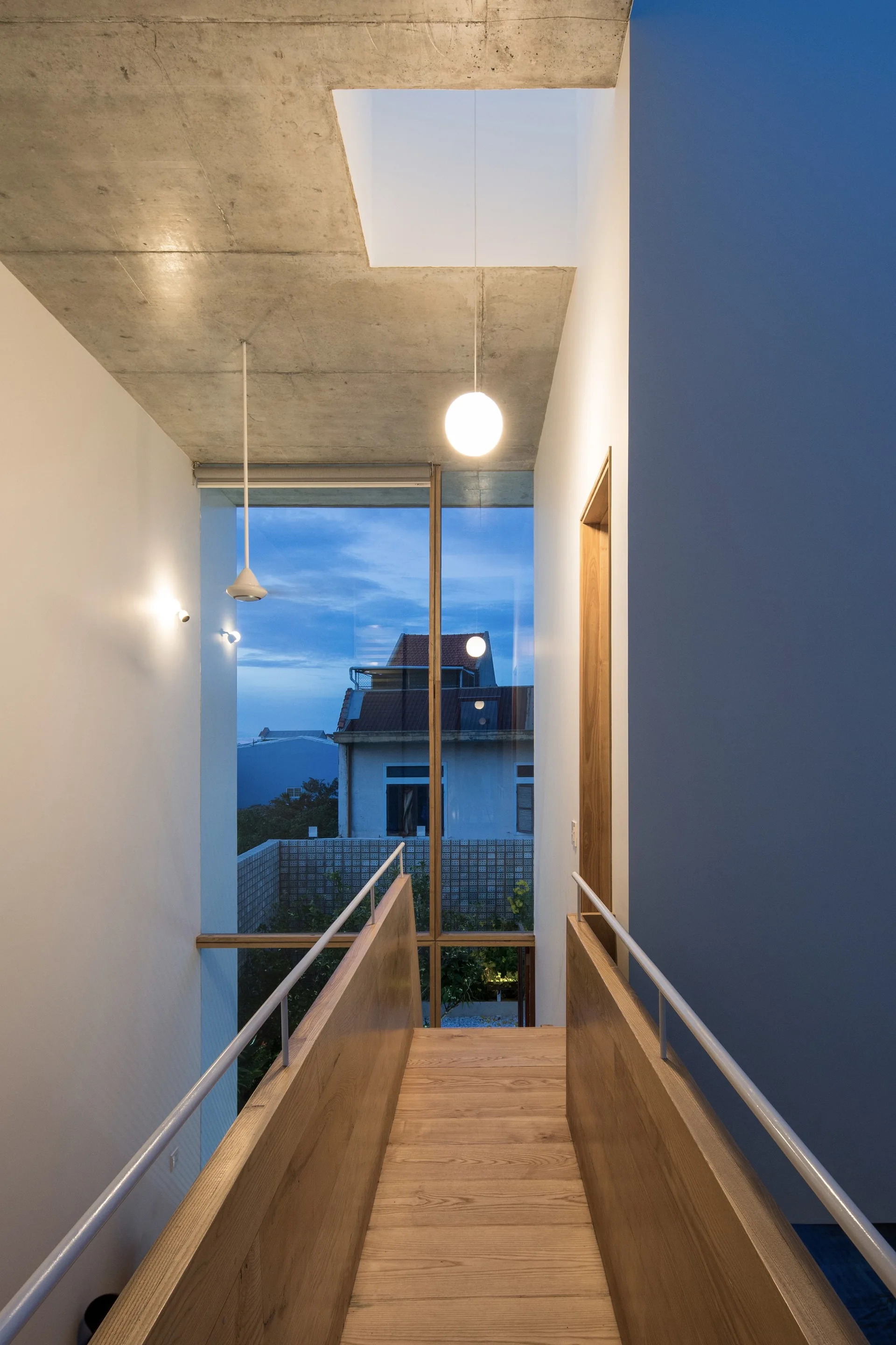 A wooden interior bridge walkway with white metal railings sits beneath a raw concrete ceiling and a glowing globe pendant light.