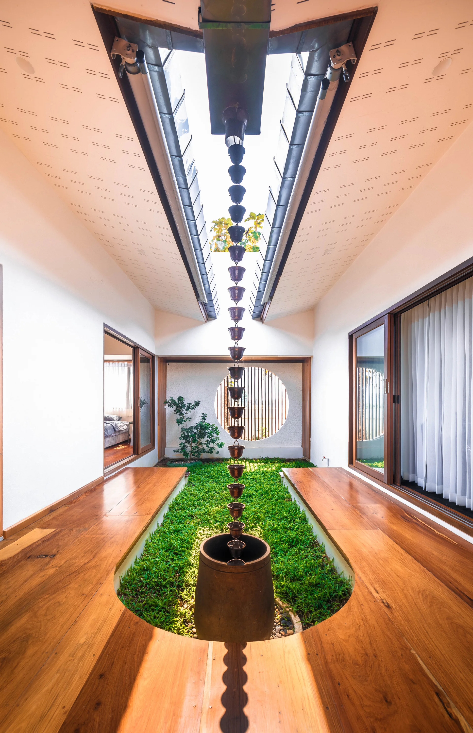 A modern indoor courtyard featuring a central strip of grass with a vertical metal rain chain, polished wooden walkways, and white walls under a skylight.