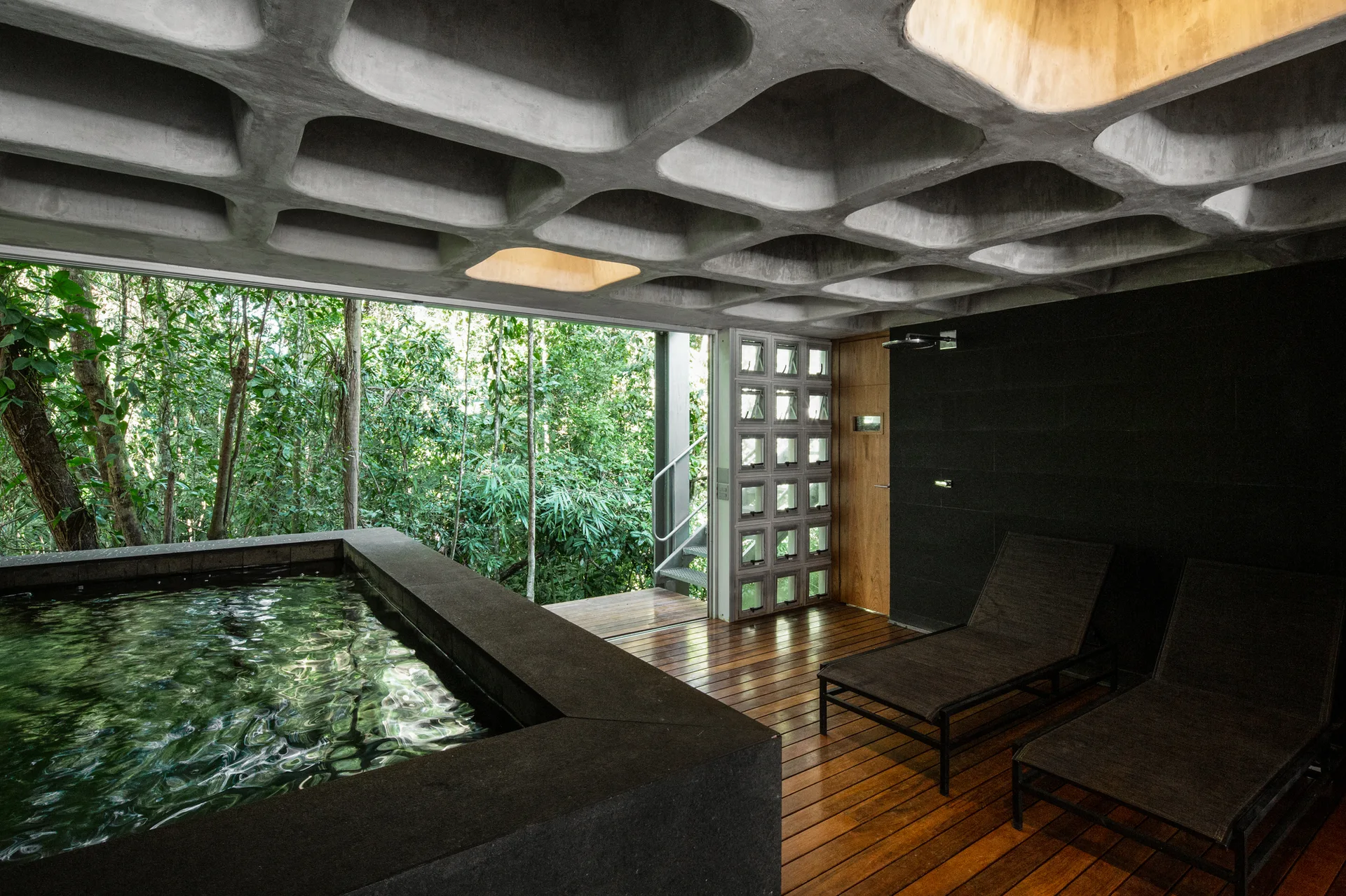 A modern indoor spa with a black plunge pool, wood flooring, and lounge chairs beneath a concrete waffle ceiling looking out at a forest.