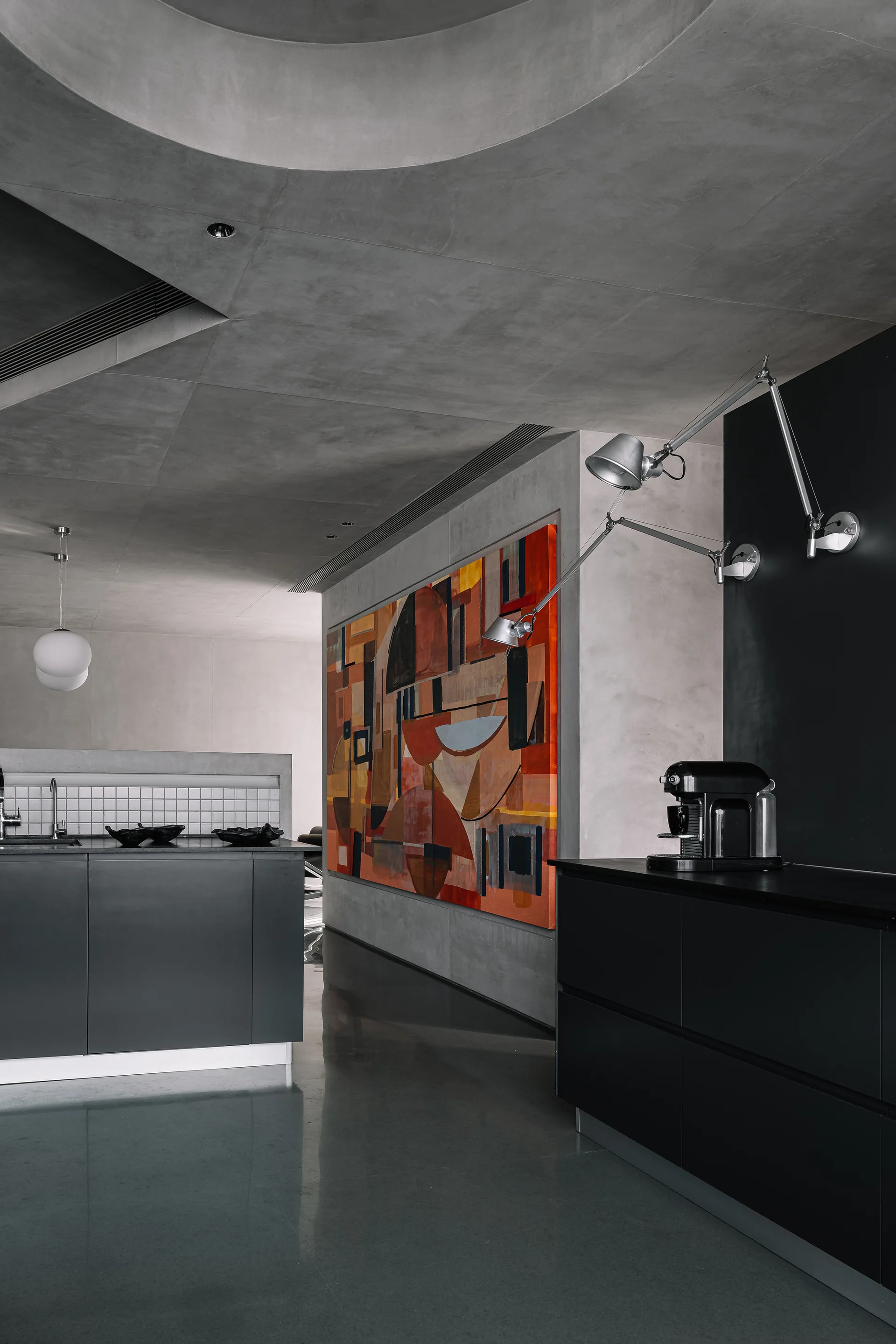 A minimalist gray concrete kitchen features matte black cabinets, an articulated wall lamp, and a large colorful abstract painting.