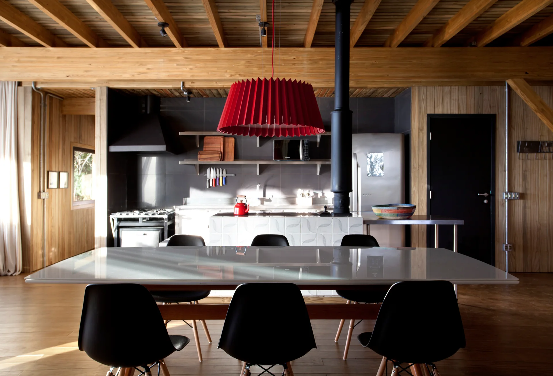 A rustic modern kitchen and dining area featuring exposed ceiling beams, a red pleated pendant light, and black dining chairs around a glossy table.