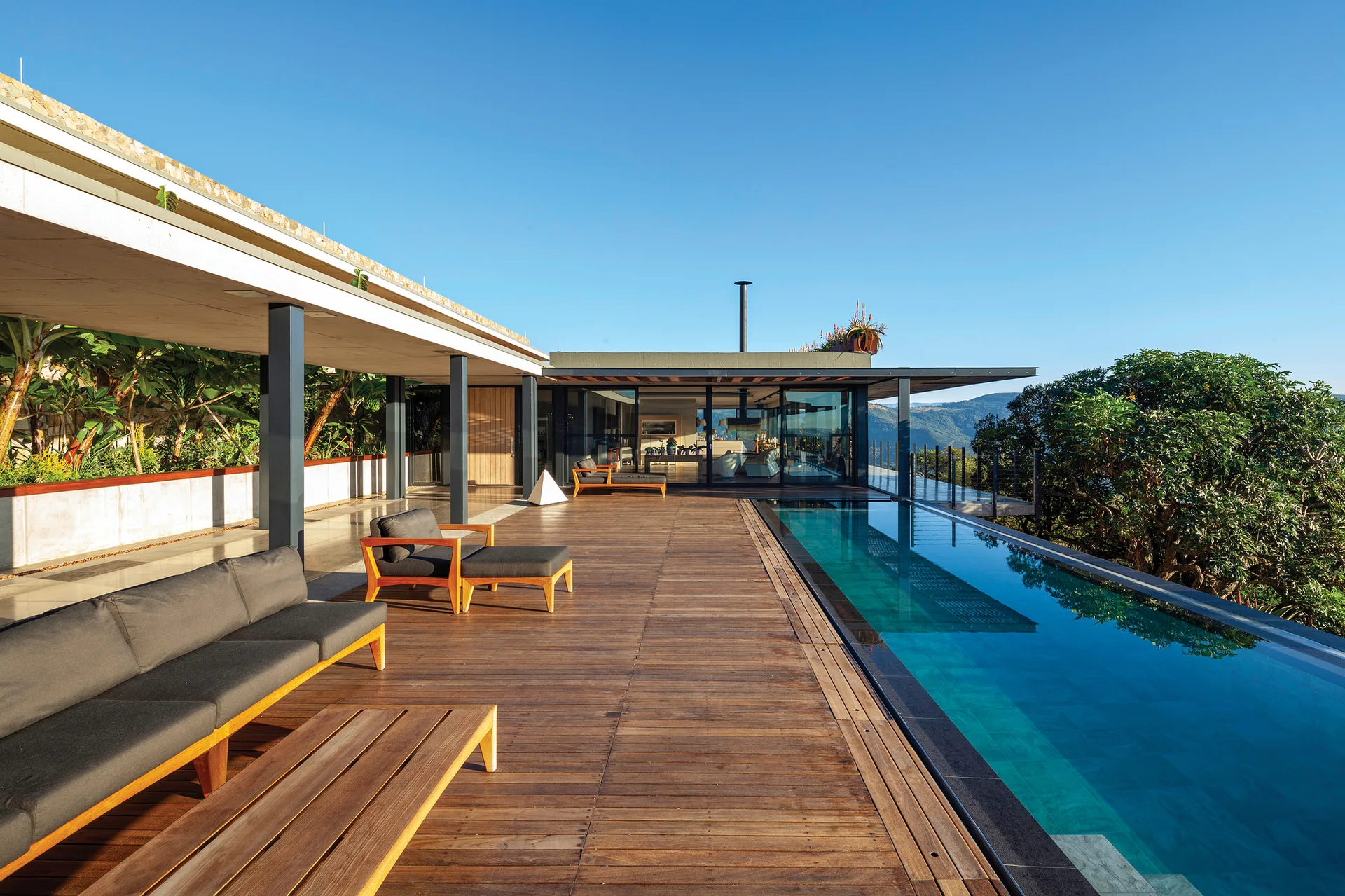 A modern wood-plank deck features a long rectangular lap pool, dark grey outdoor sofas, and a concrete roof overhang under a clear sky.