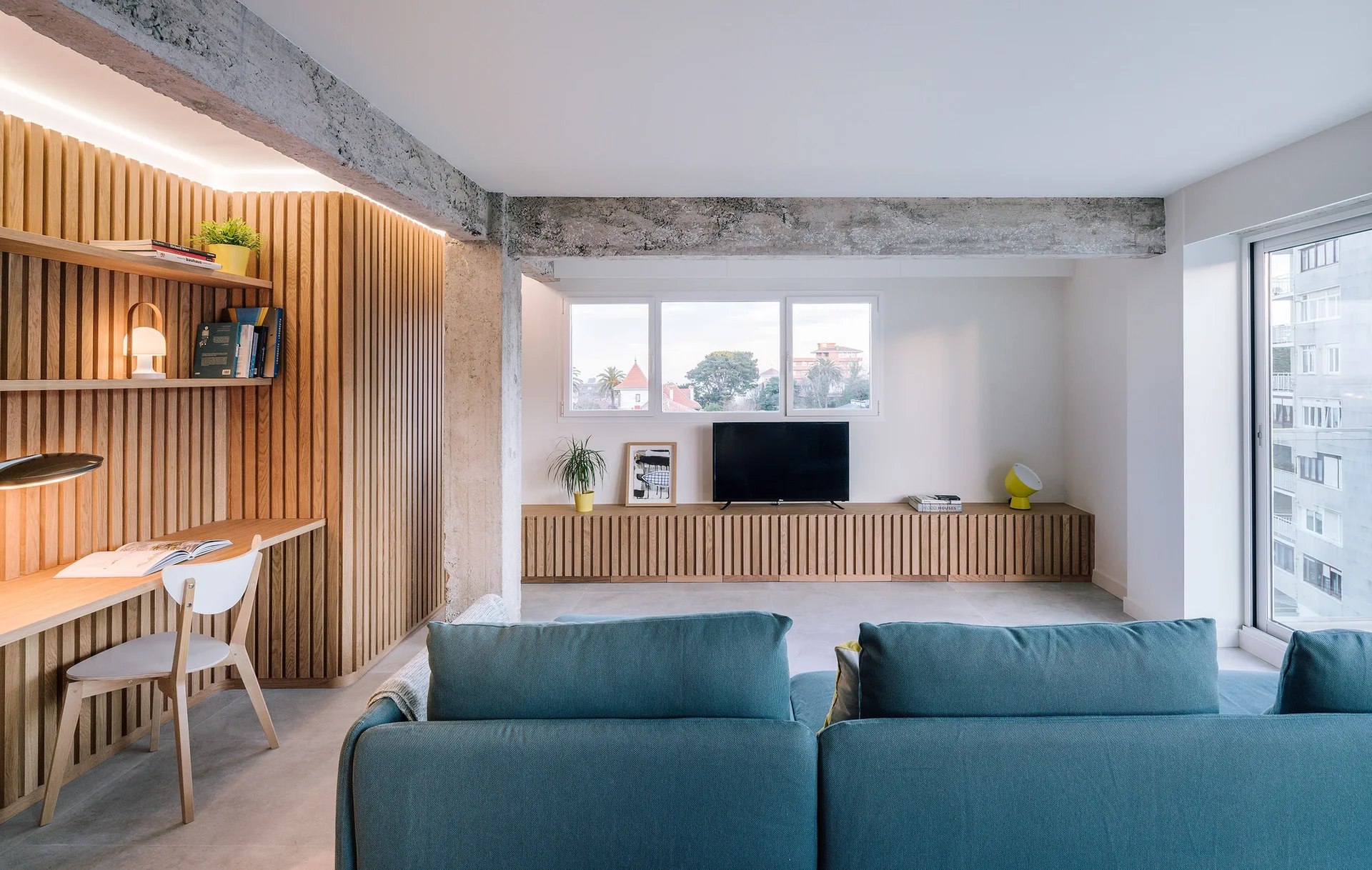 A modern living room features a teal sofa, a wood slatted wall with an integrated desk, and an exposed concrete ceiling beam.