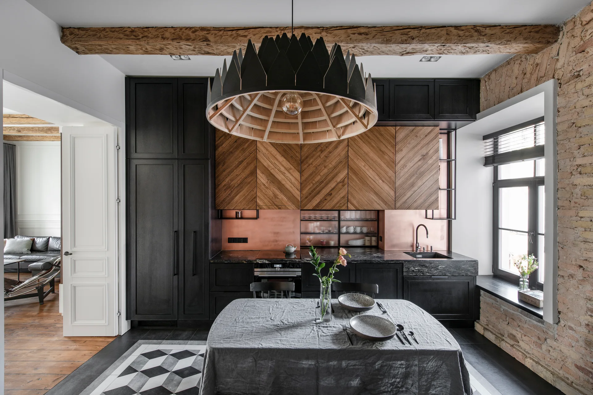 A modern kitchen features black cabinetry, chevron-patterned wood upper cabinets, a warm copper backsplash, and an exposed brick wall near a window.