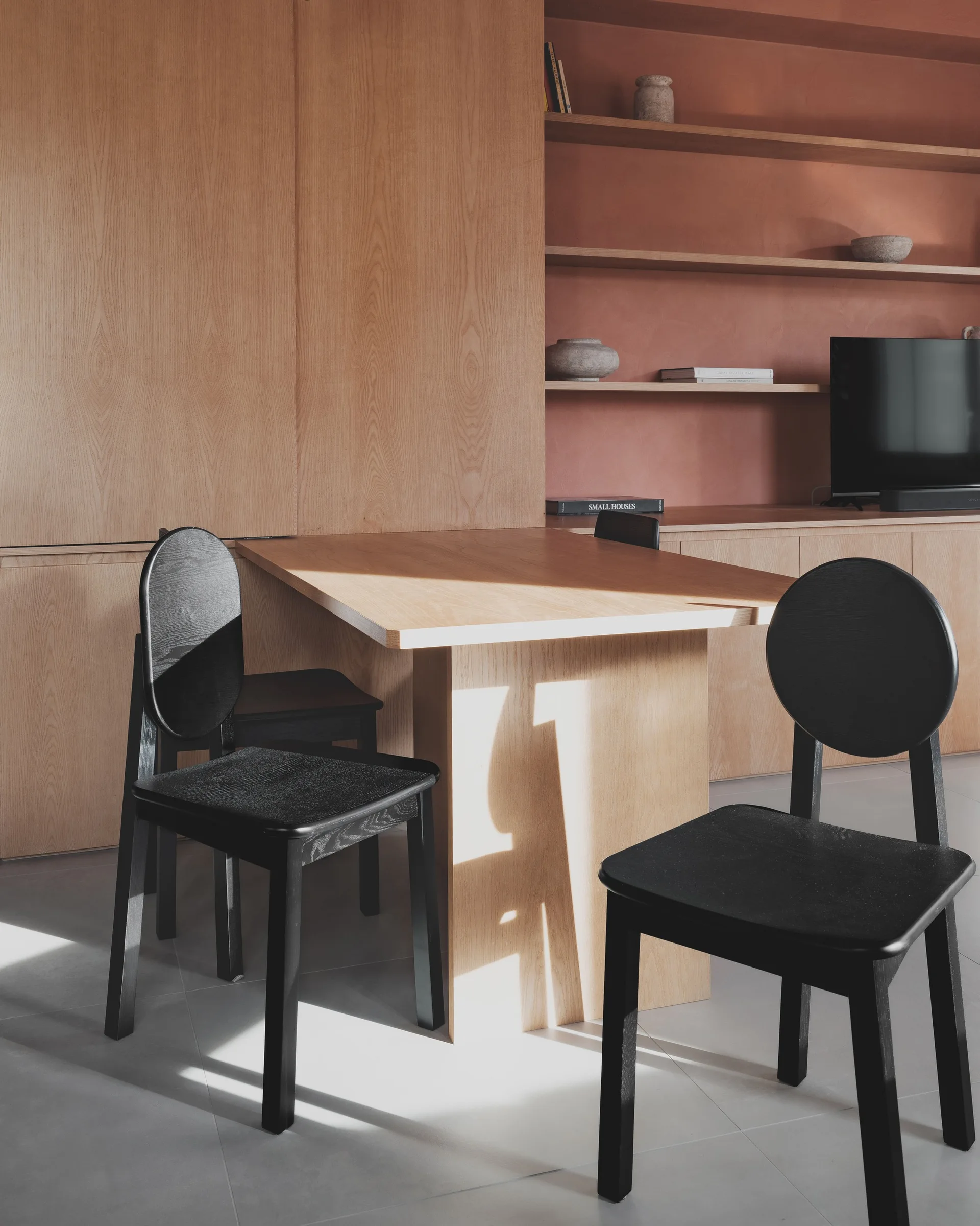 A minimalist dining area with a light oak built-in table, two black wooden chairs, and a terracotta-colored recessed wall niche with shelves.