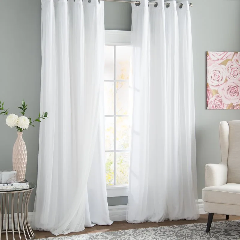 Layered white curtains hang over a window next to a beige wingback chair and a metal side table with a vase.