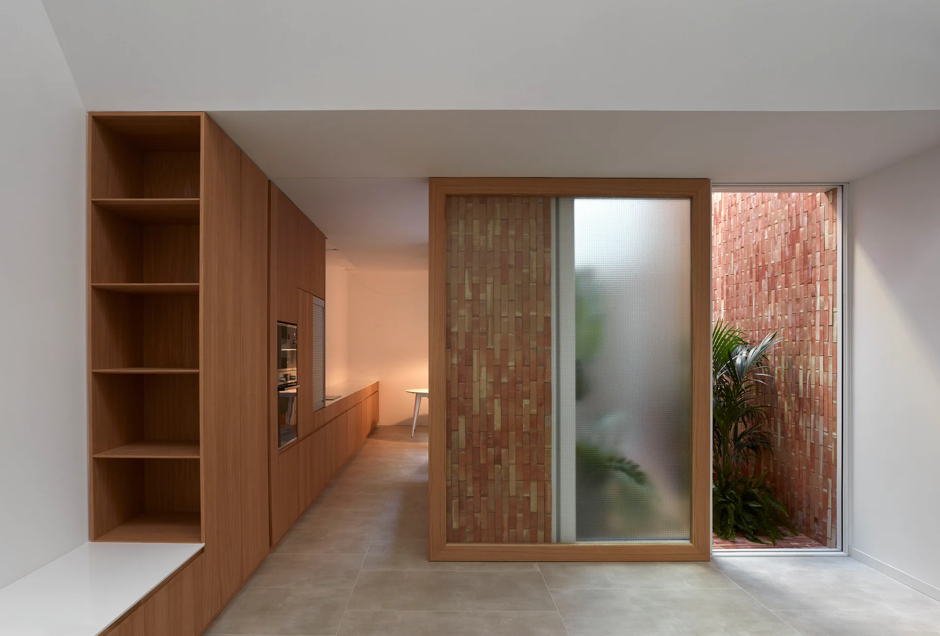 A minimalist modern kitchen featuring warm flat-front wood cabinetry, beige tile flooring, and a partition made of textured glass and vertical brick.