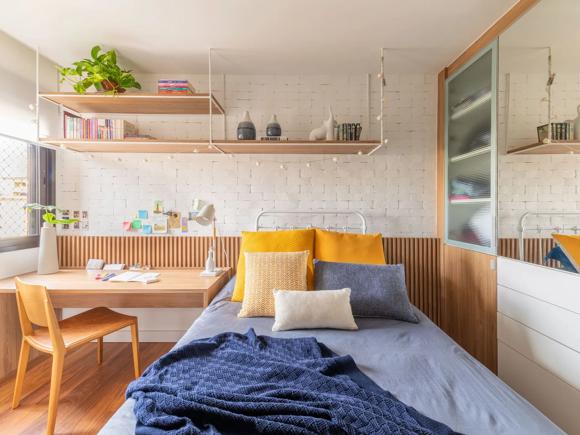 A compact bedroom features a bed with yellow and blue pillows next to an integrated wooden desk against a white brick wall.