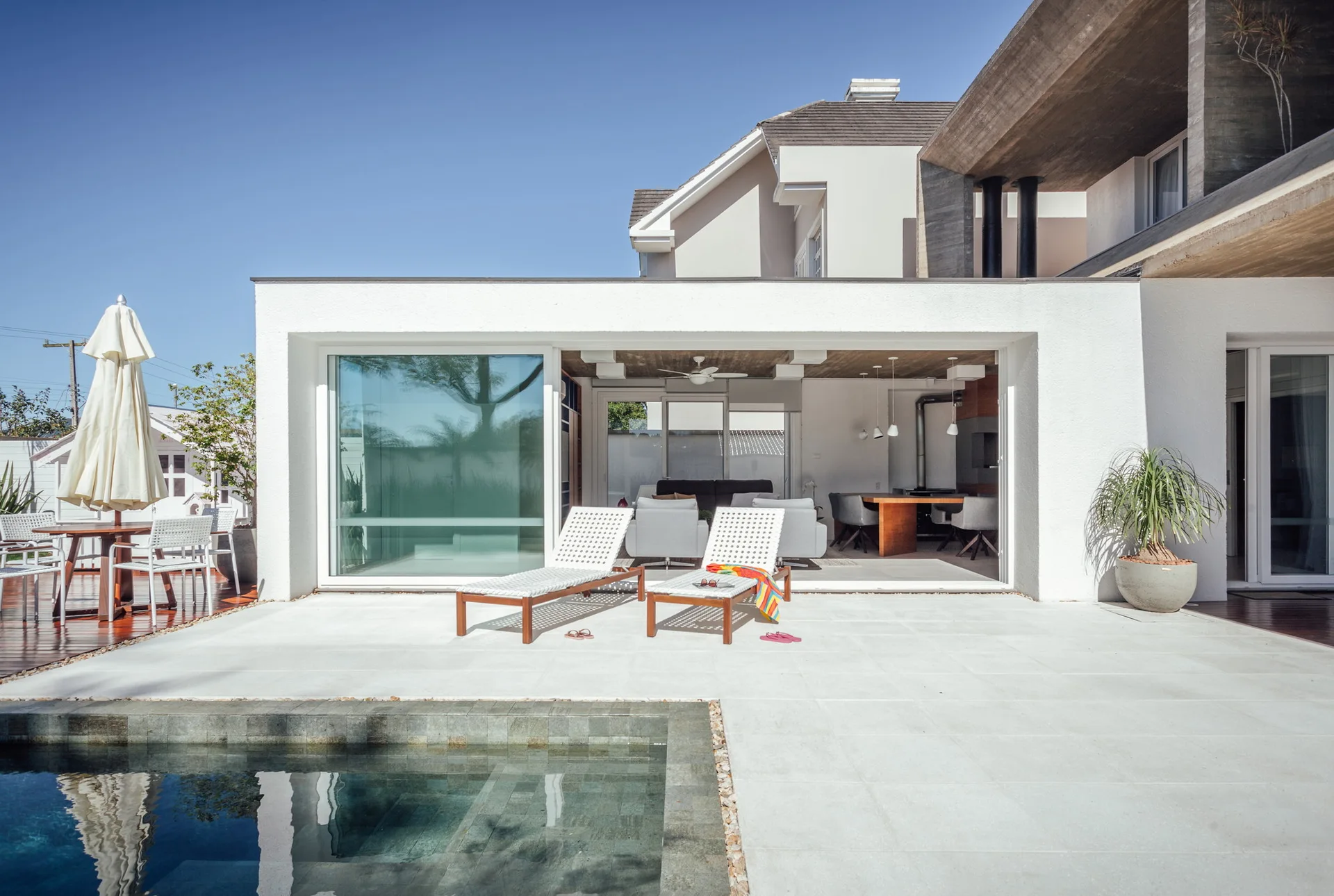 A modern poolside patio with two woven sun loungers sits next to an open-concept living and dining area with large glass sliding doors.