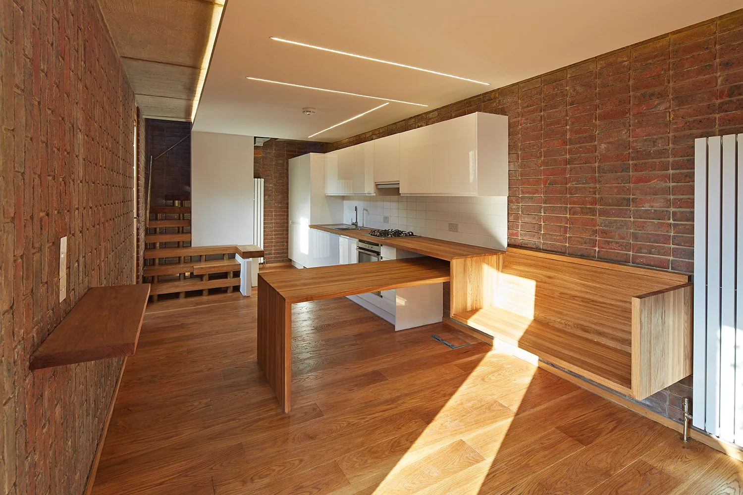 A modern kitchen featuring white glossy upper cabinets, exposed red brick walls, wood flooring, and an integrated wooden dining table and bench.