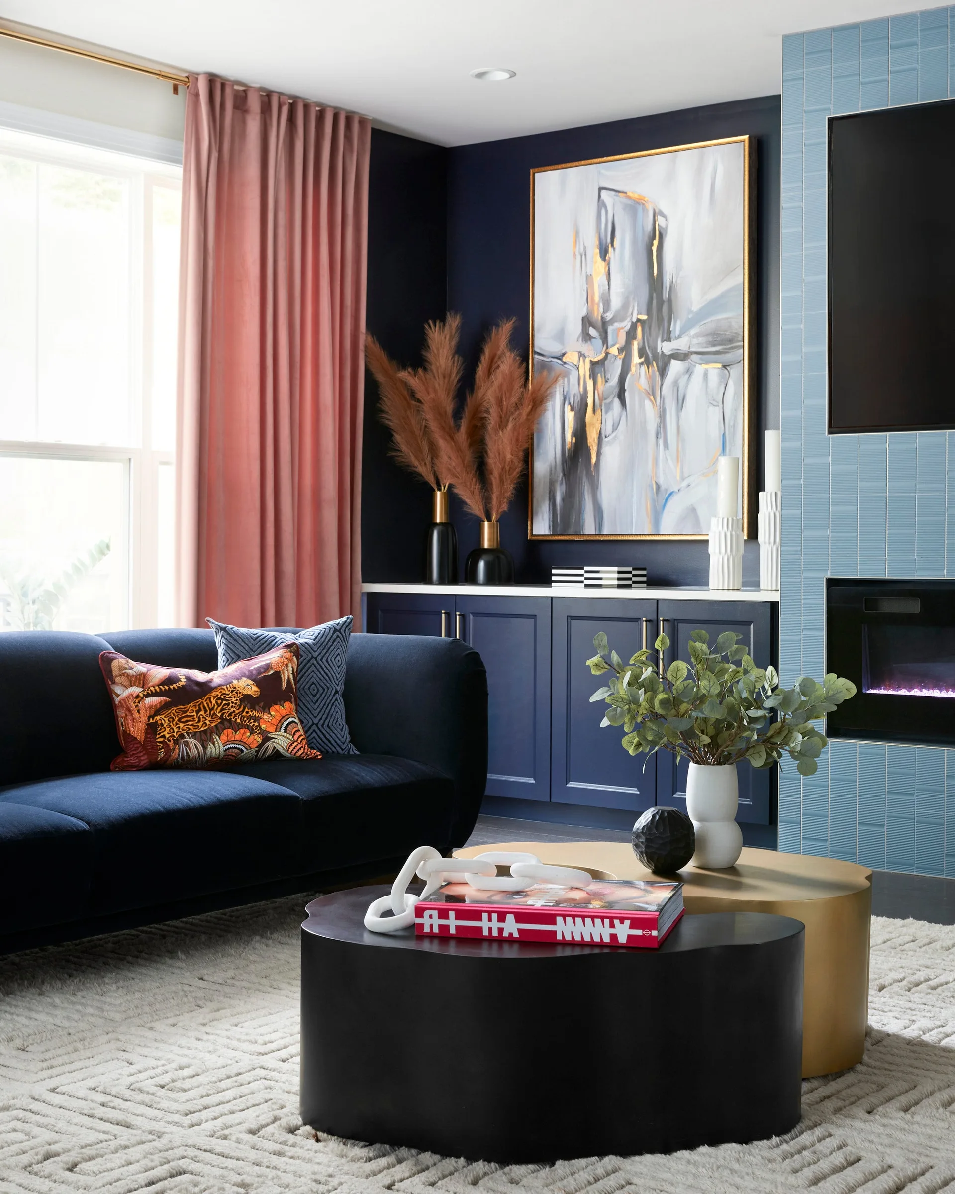 A dark navy living room features a blue velvet sofa, blush pink velvet curtains, a blue tiled fireplace, and nested black and gold coffee tables.
