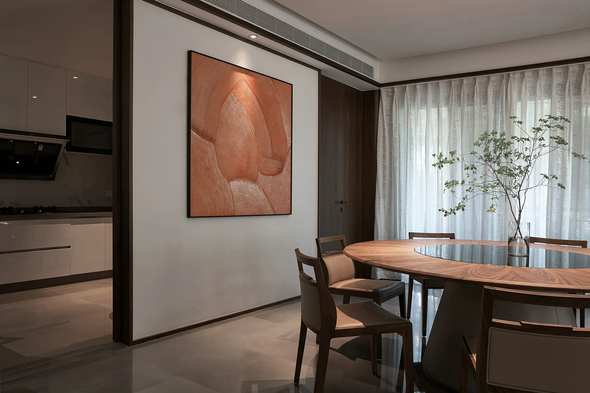 A modern dining room features a round wooden table, chairs, sheer white curtains, and a large textured copper painting on a white partition wall.
