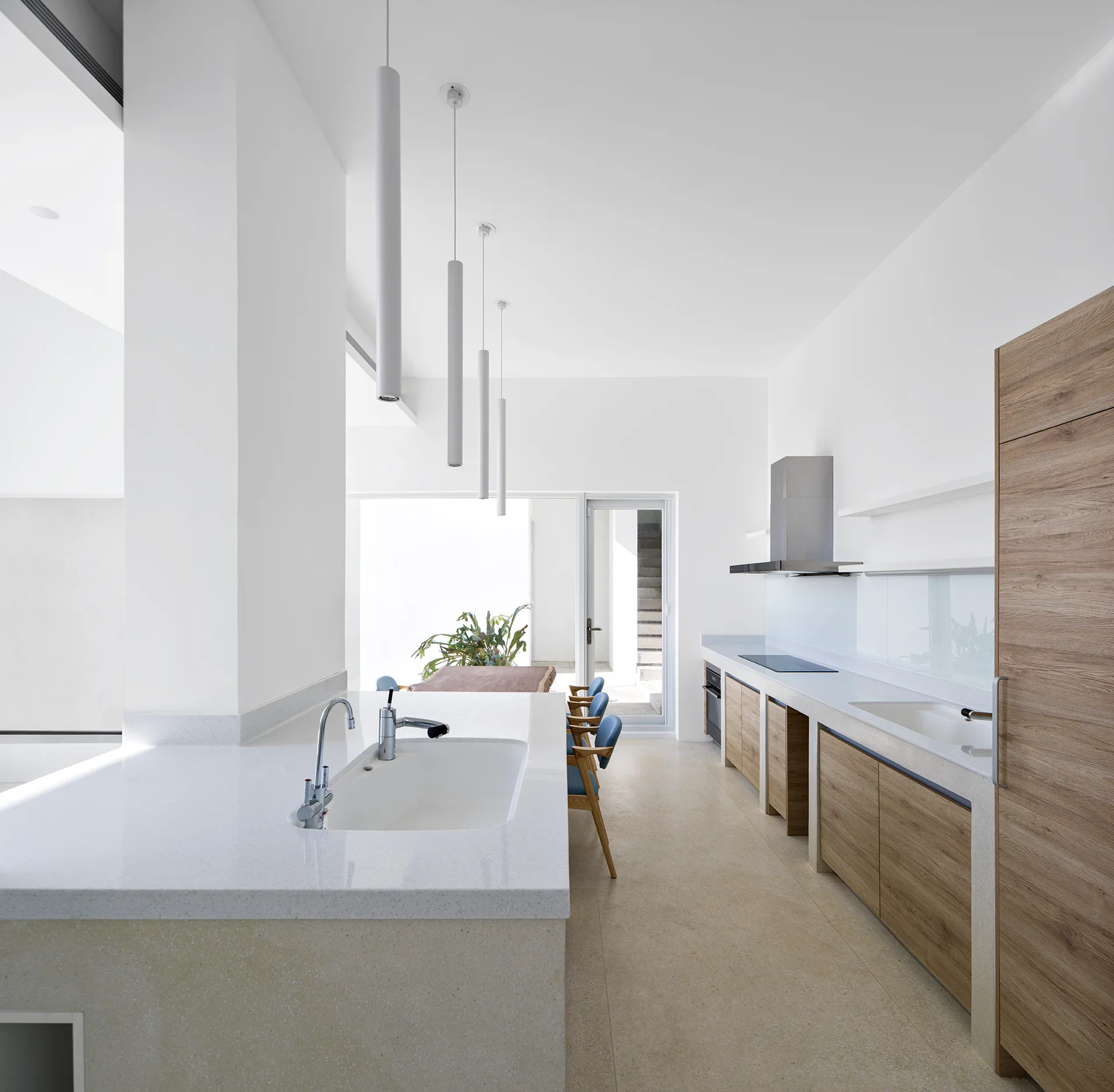 A bright minimalist kitchen features a white stone island, light wood cabinets, and three slender white cylindrical pendant lights.
