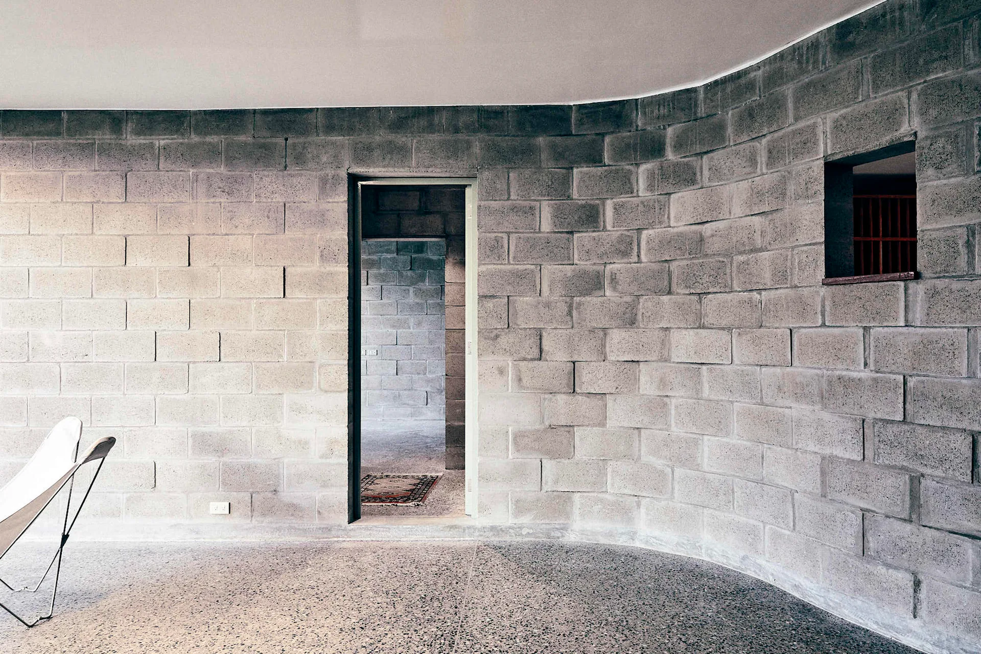 A curved wall made of gray concrete blocks stands in an industrial interior with polished terrazzo flooring and a white butterfly chair.