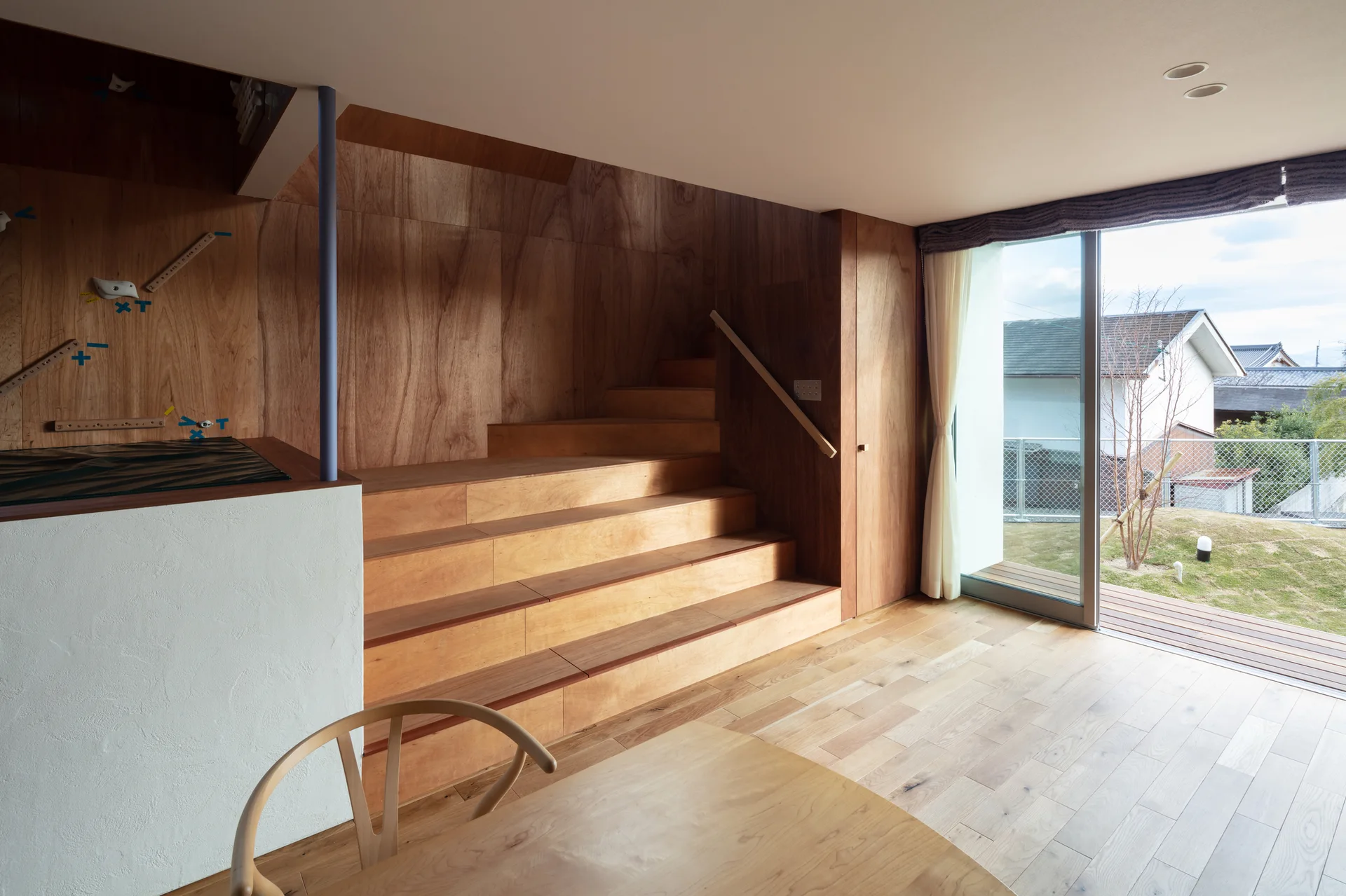 A light-filled living area features a wide wooden staircase, wood-paneled walls, and a large sliding glass door opening to a grassy yard.