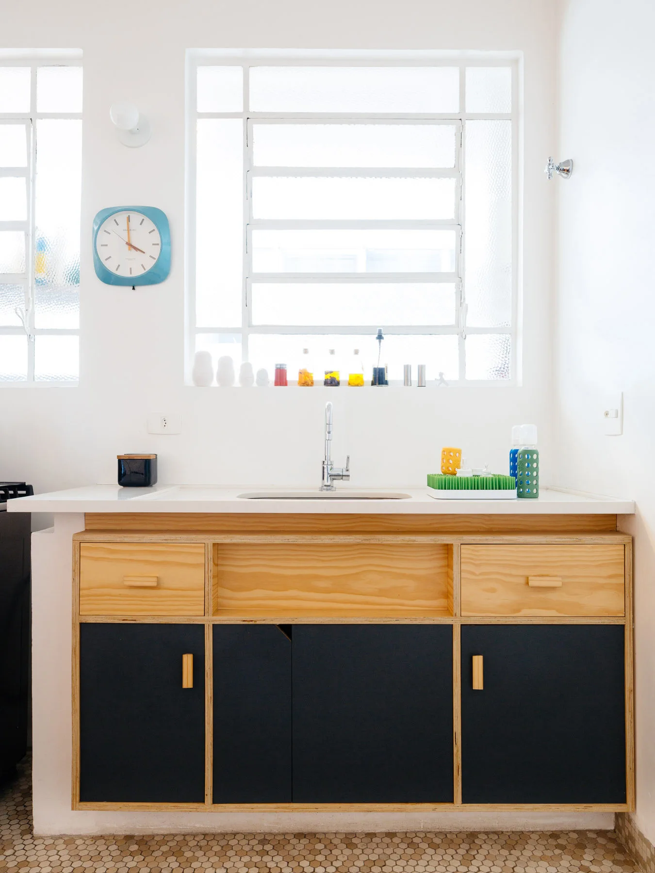 A minimalist kitchen sink features two-tone plywood cabinetry with dark blue doors below large multi-pane windows and a hexagonal tile floor.