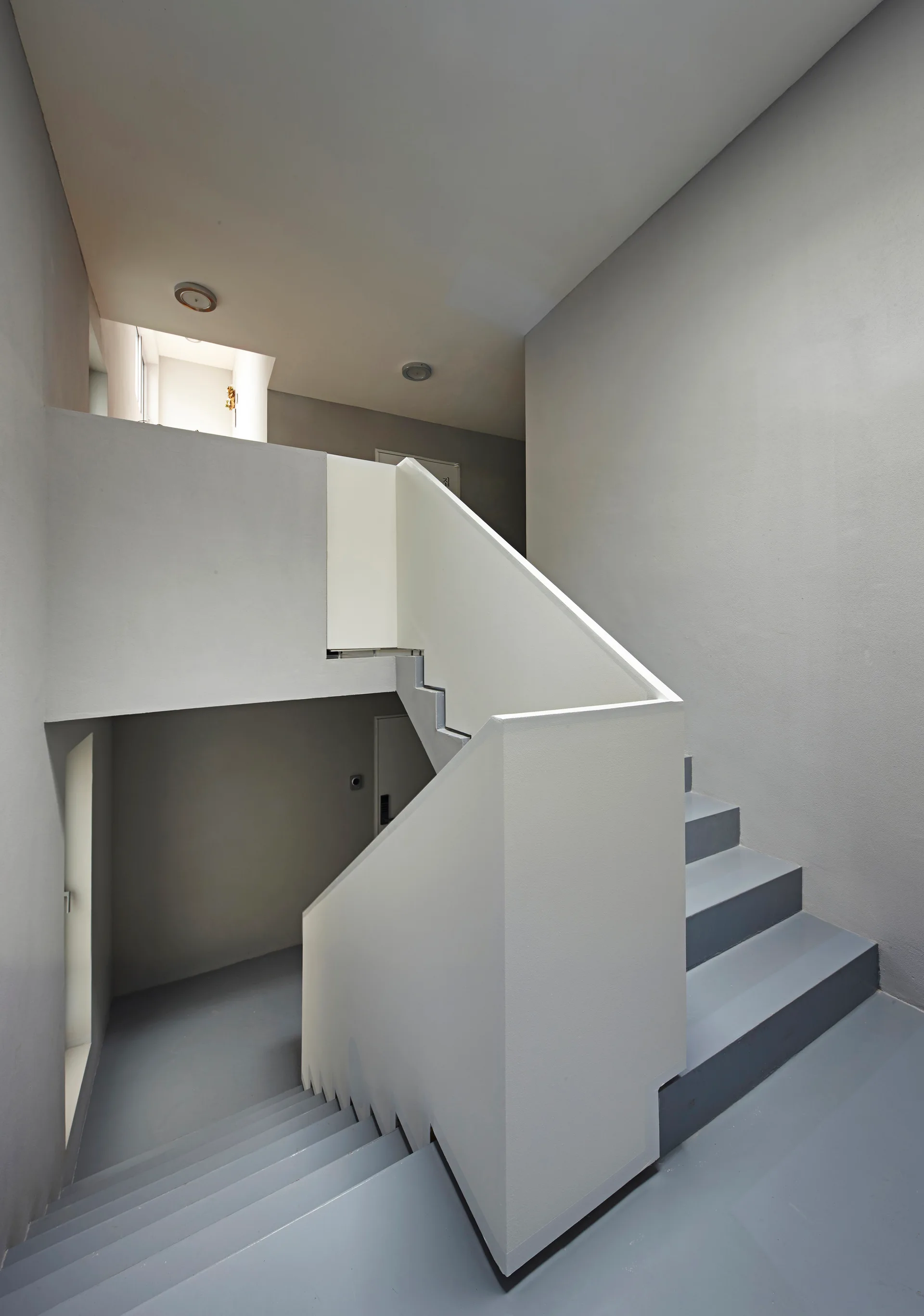 A minimalist staircase features matte grey painted steps, a solid white block railing, and smooth grey walls with flush ceiling lights.
