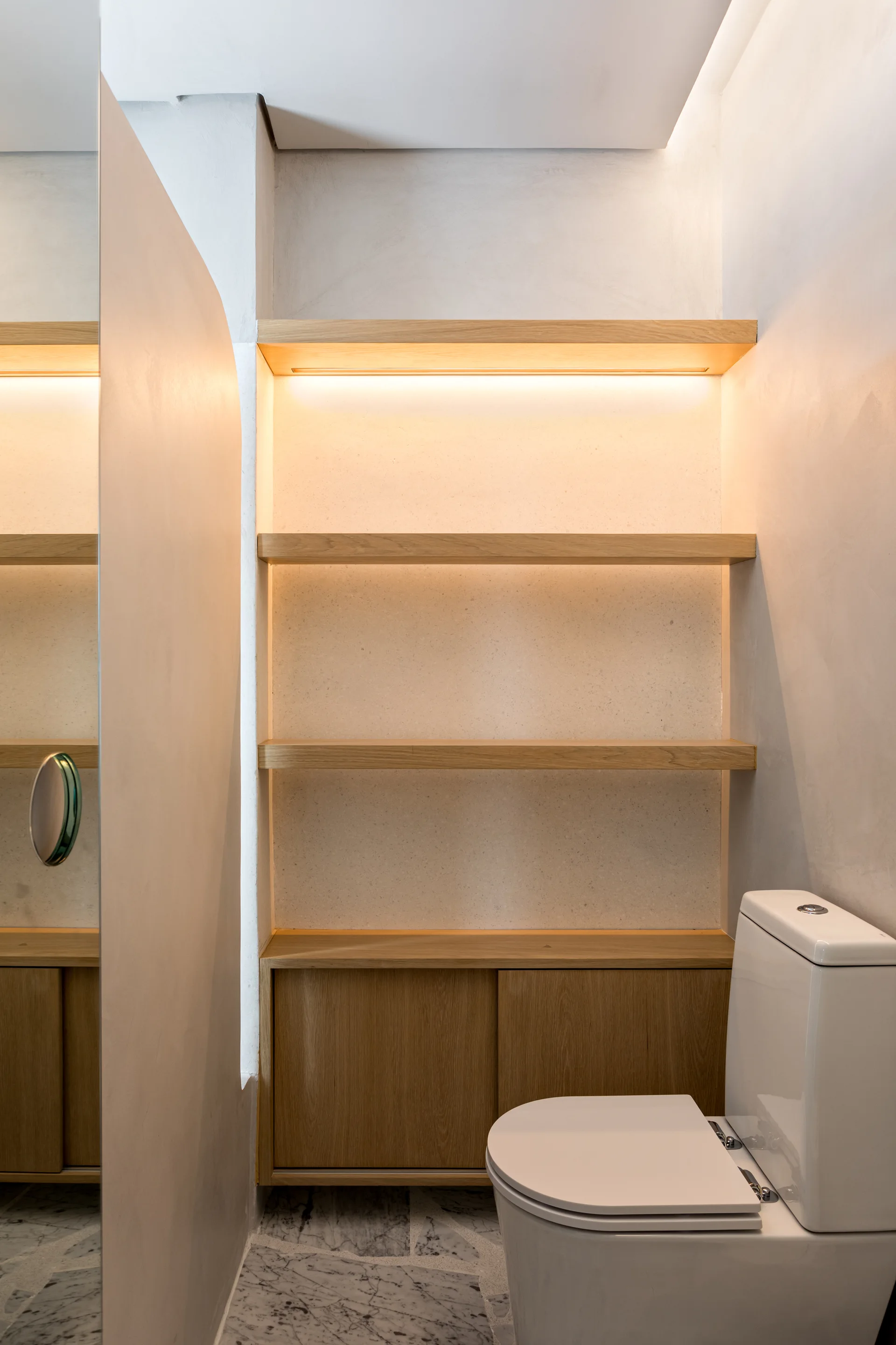 A minimalist bathroom features light wood built-in shelving with integrated warm lighting, a lower cabinet, marble flooring, and a white toilet.